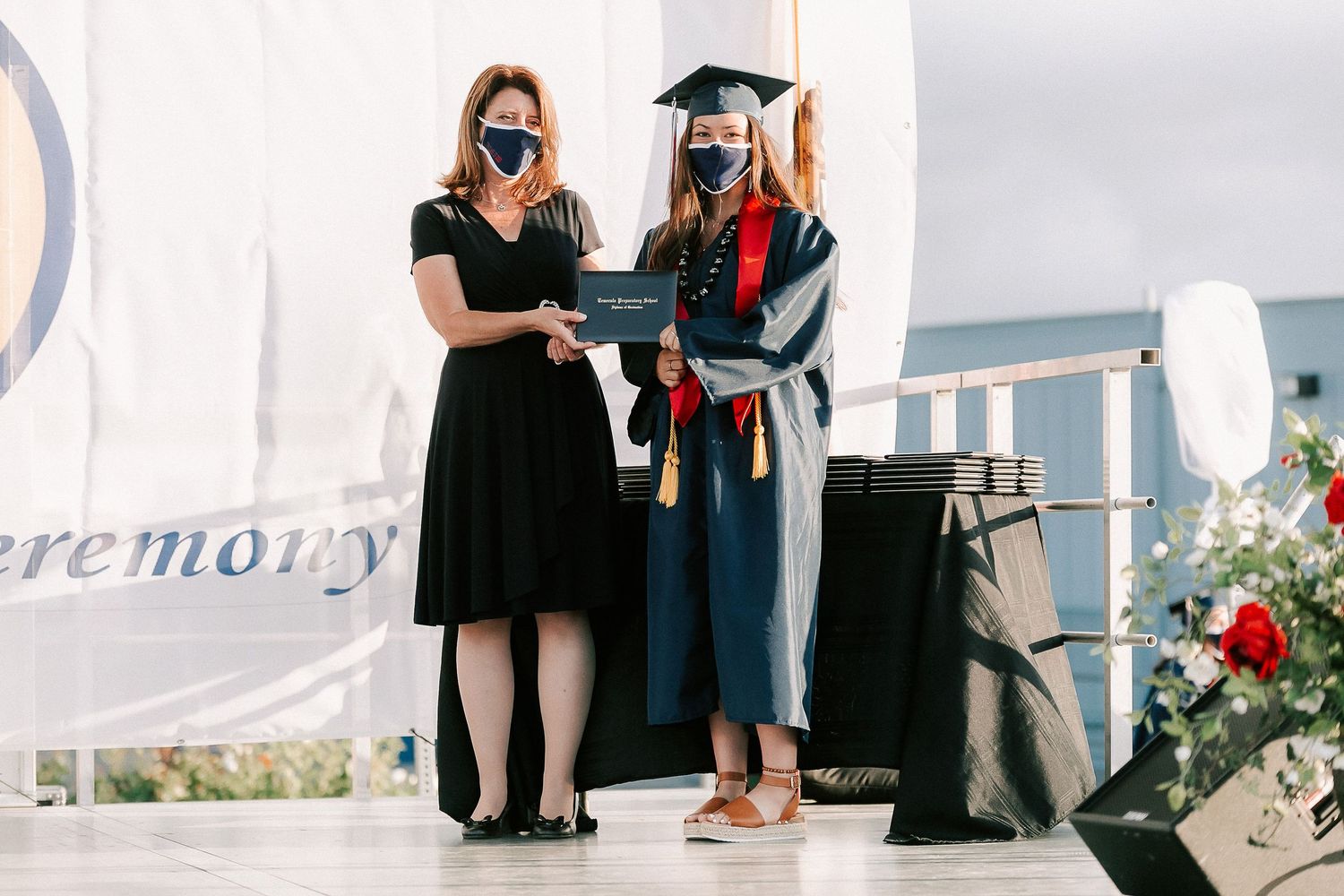 Graduate in cap and gown holding diploma with presenter in black dress at 2020 graduation ceremony.