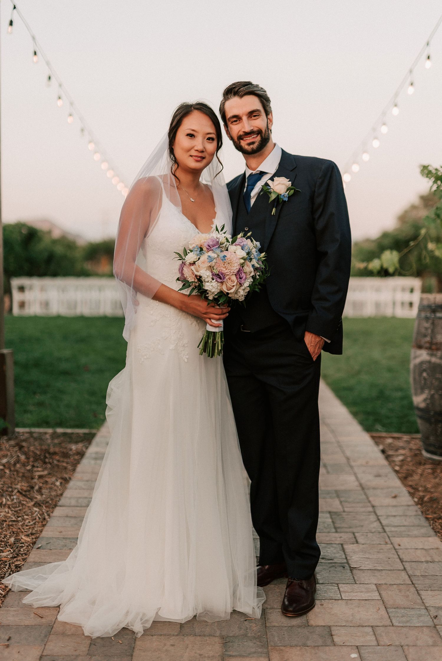 Bride in white gown with veil holding floral bouquet stands beside groom in navy suit on outdoor wedding venue path.