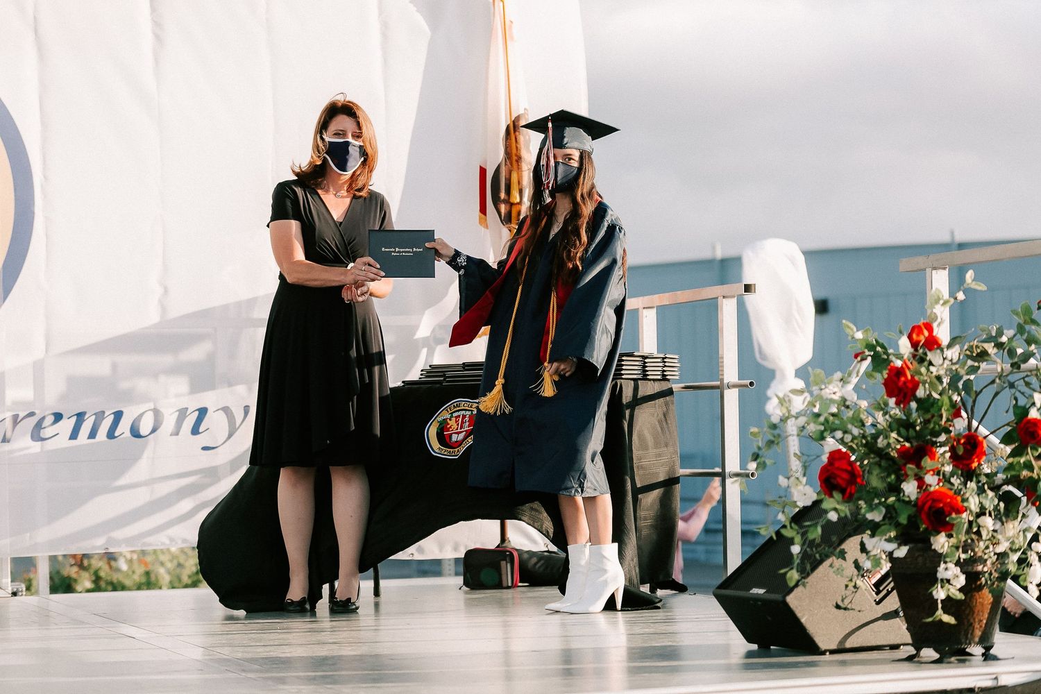 Graduate receiving diploma on stage at outdoor graduation ceremony, both wearing masks, with red roses nearby.
