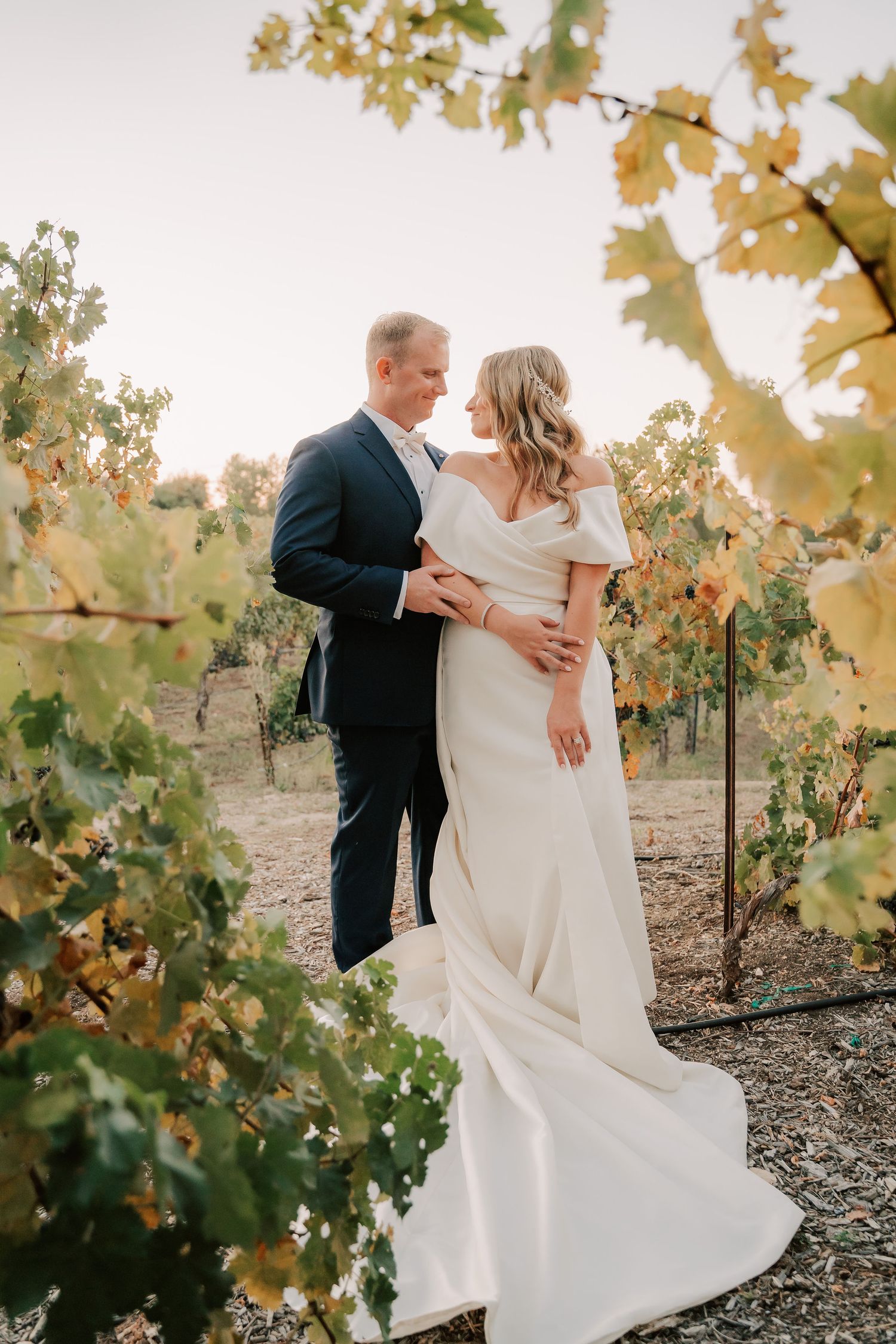 Bride and groom embrace in a sunlit vineyard, she in a white off-shoulder gown, he in a navy suit.