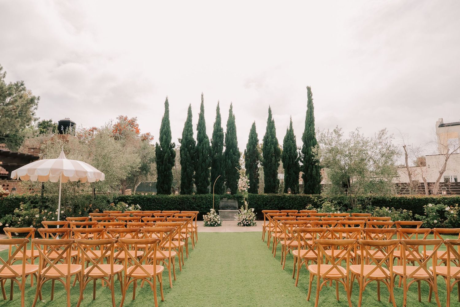 Outdoor wedding ceremony setup with wooden chairs on green lawn, tall cypress trees, and white umbrella at elegant garden venue.