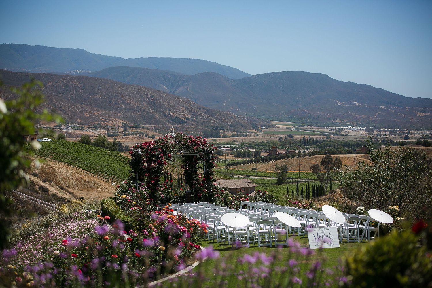 Outdoor wedding ceremony setup with white chairs overlooking a scenic vineyard valley and mountain landscape under clear blue skies.