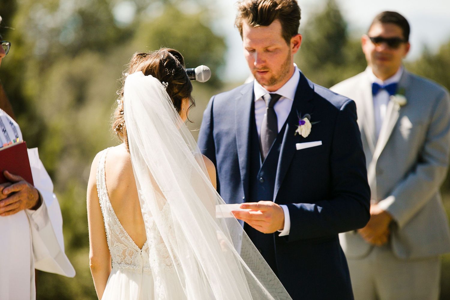 Groom reads wedding vows to bride in white dress and veil during outdoor wedding ceremony.