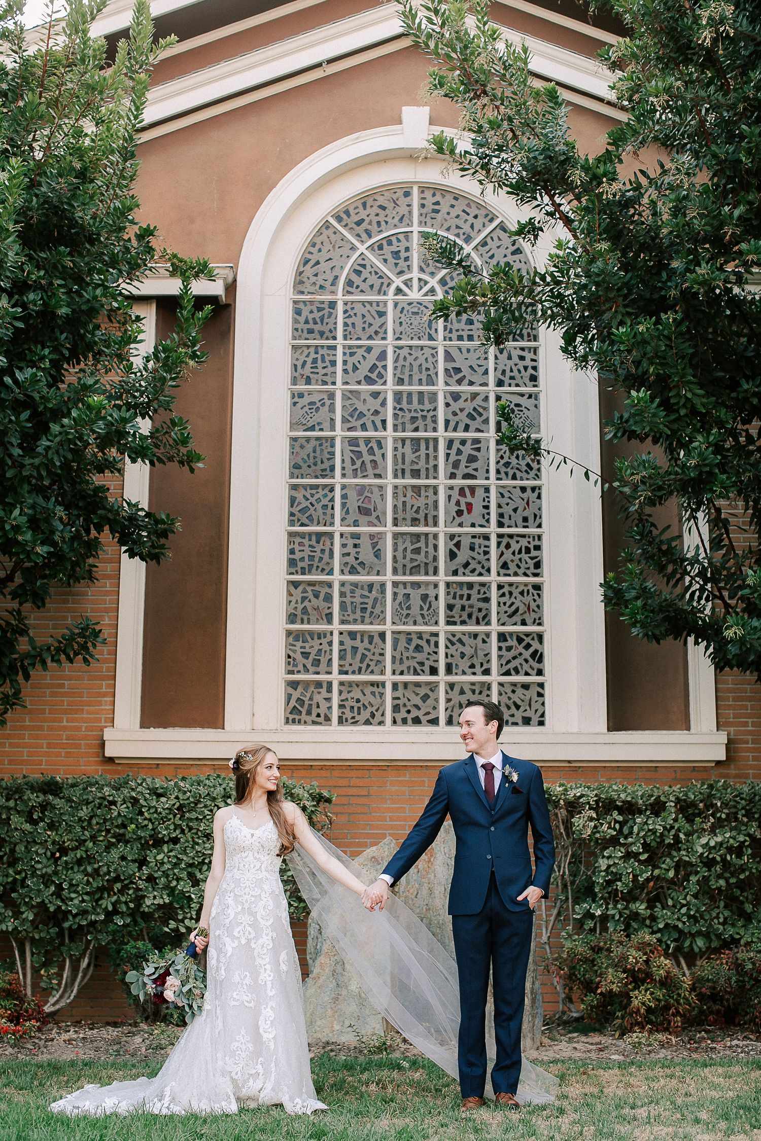 Bride and groom hold hands outside a brick church with an arched stained glass window, surrounded by lush green trees.
