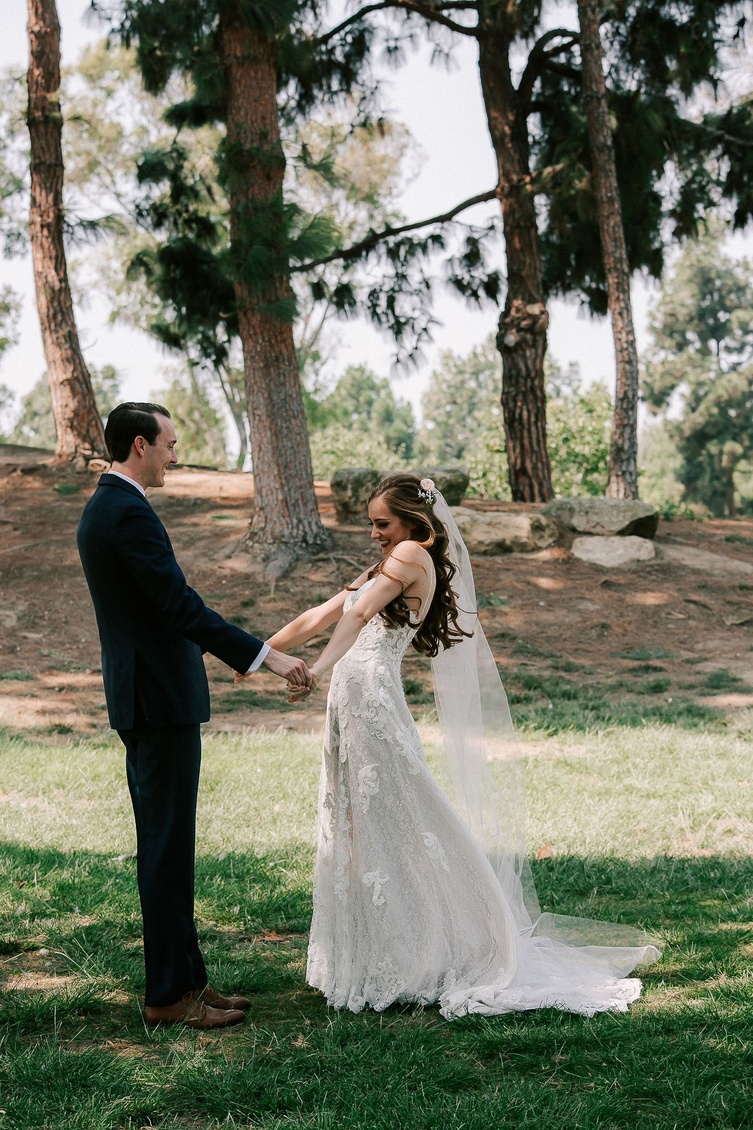 A bride and groom hold hands outdoors under tall pine trees, the bride wearing a lace gown with veil.