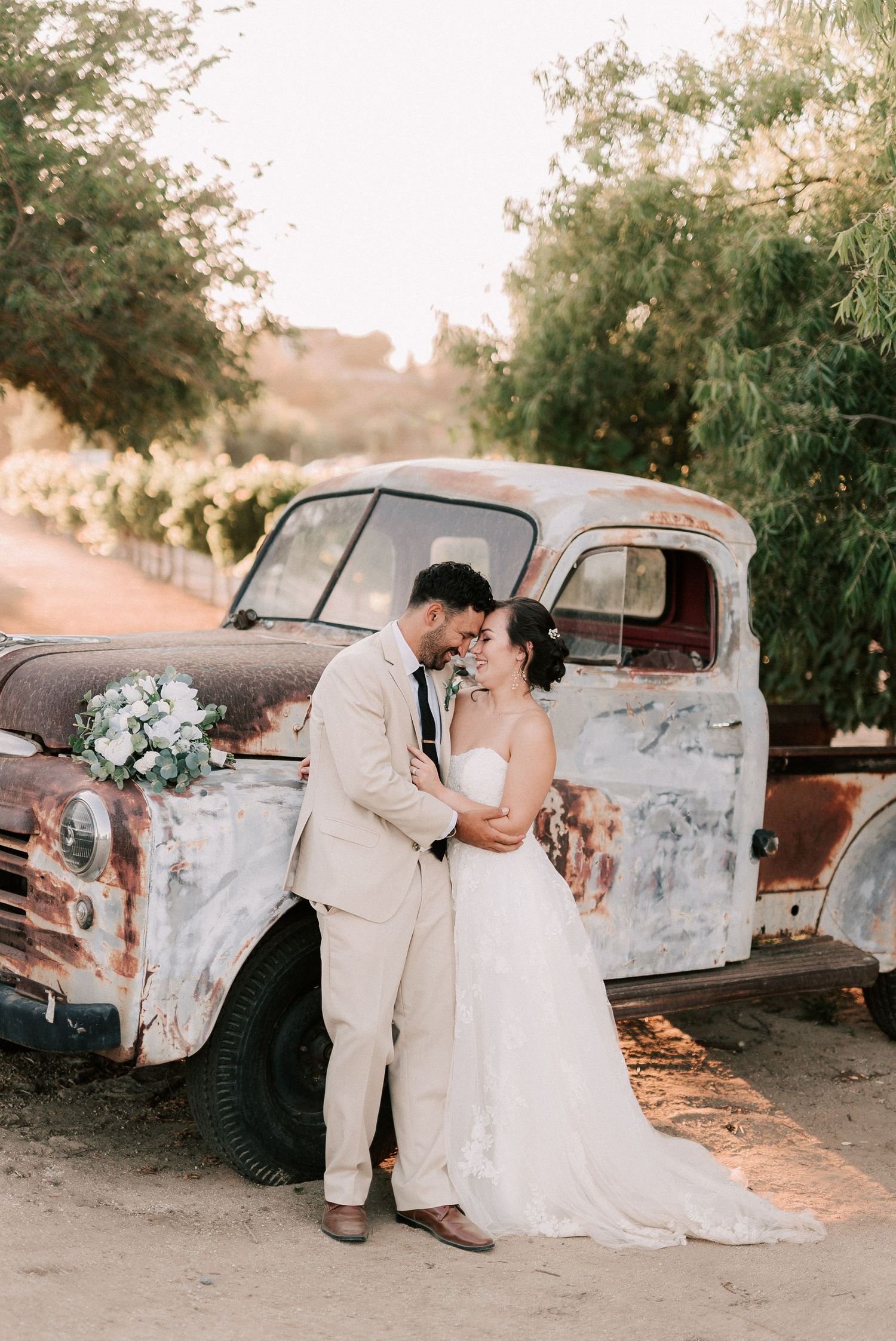 Bride and groom share a romantic moment leaning foreheads together beside a rustic vintage truck at a vineyard wedding.
