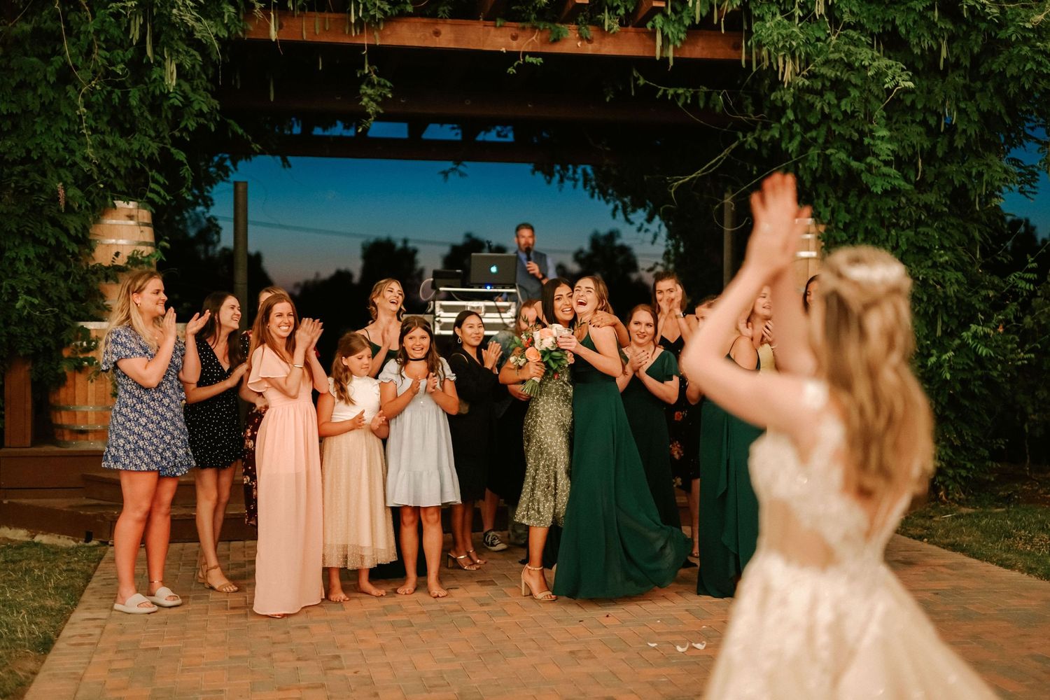Bride tossing bouquet to excited bridesmaids and guests at outdoor wedding reception under vine-covered pergola.