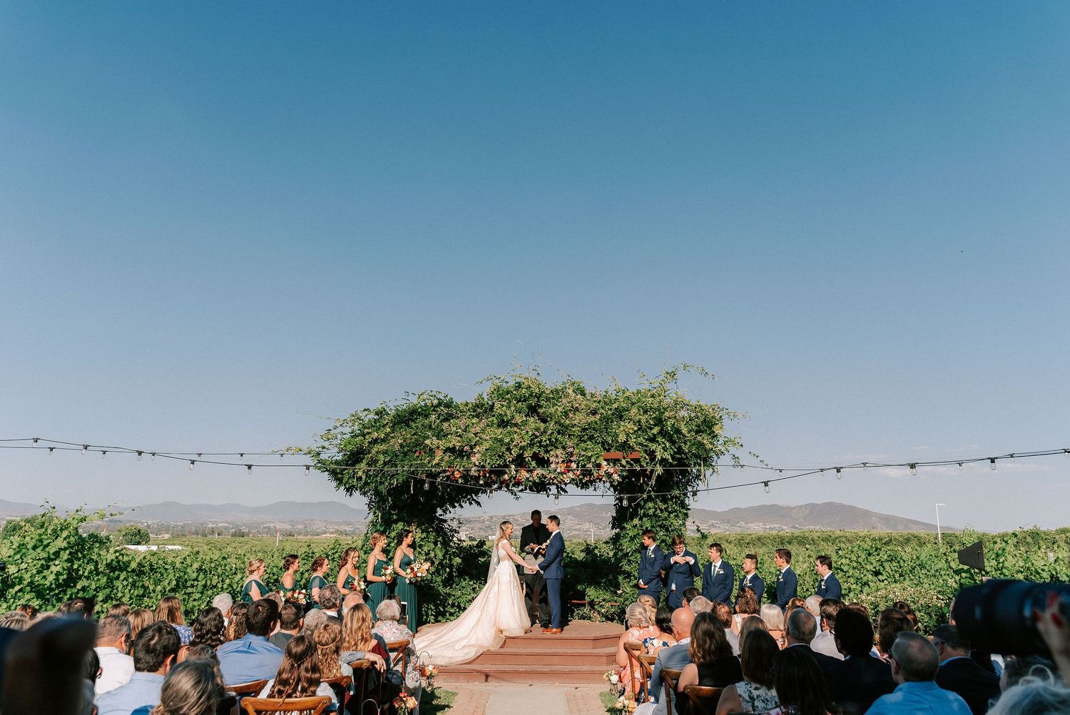 Outdoor vineyard wedding ceremony with bride and groom at floral arch, string lights, wedding party, and guests under clear blue sky.