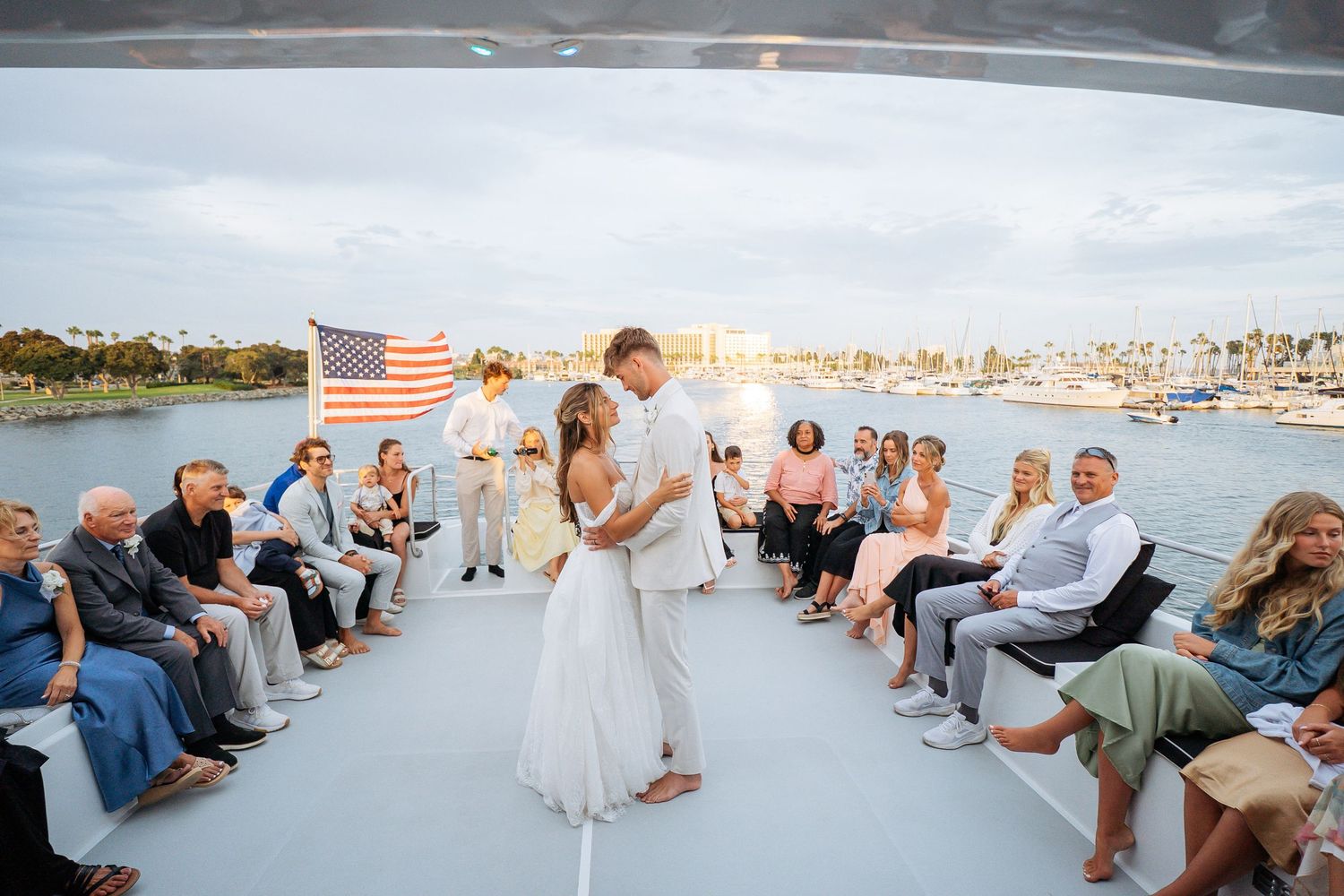 Bride and groom share first dance on yacht deck surrounded by seated wedding guests with American flag and harbor backdrop.