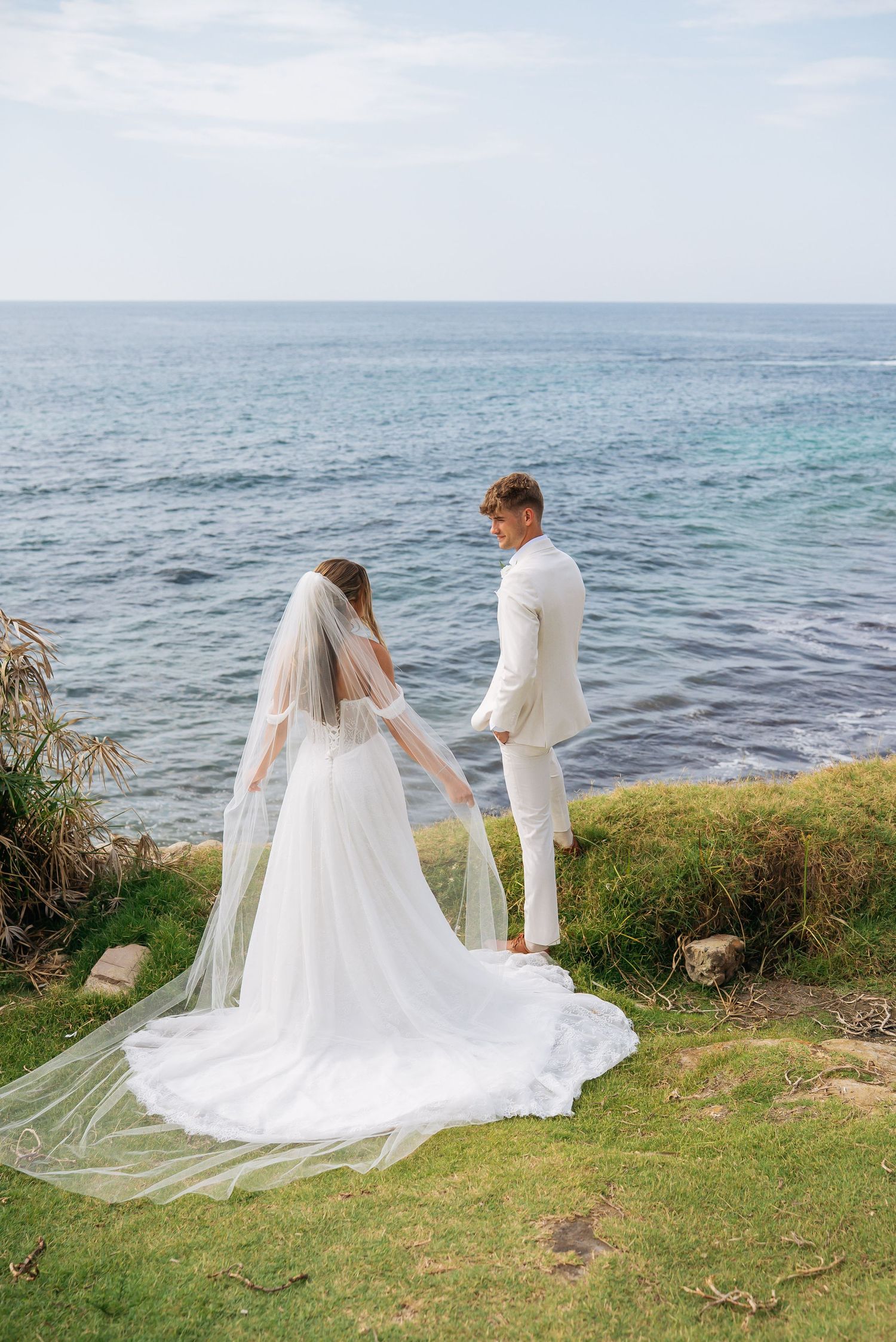 Newlywed couple holds hands while standing on a grassy seaside cliff, bride's veil flowing in the breeze.