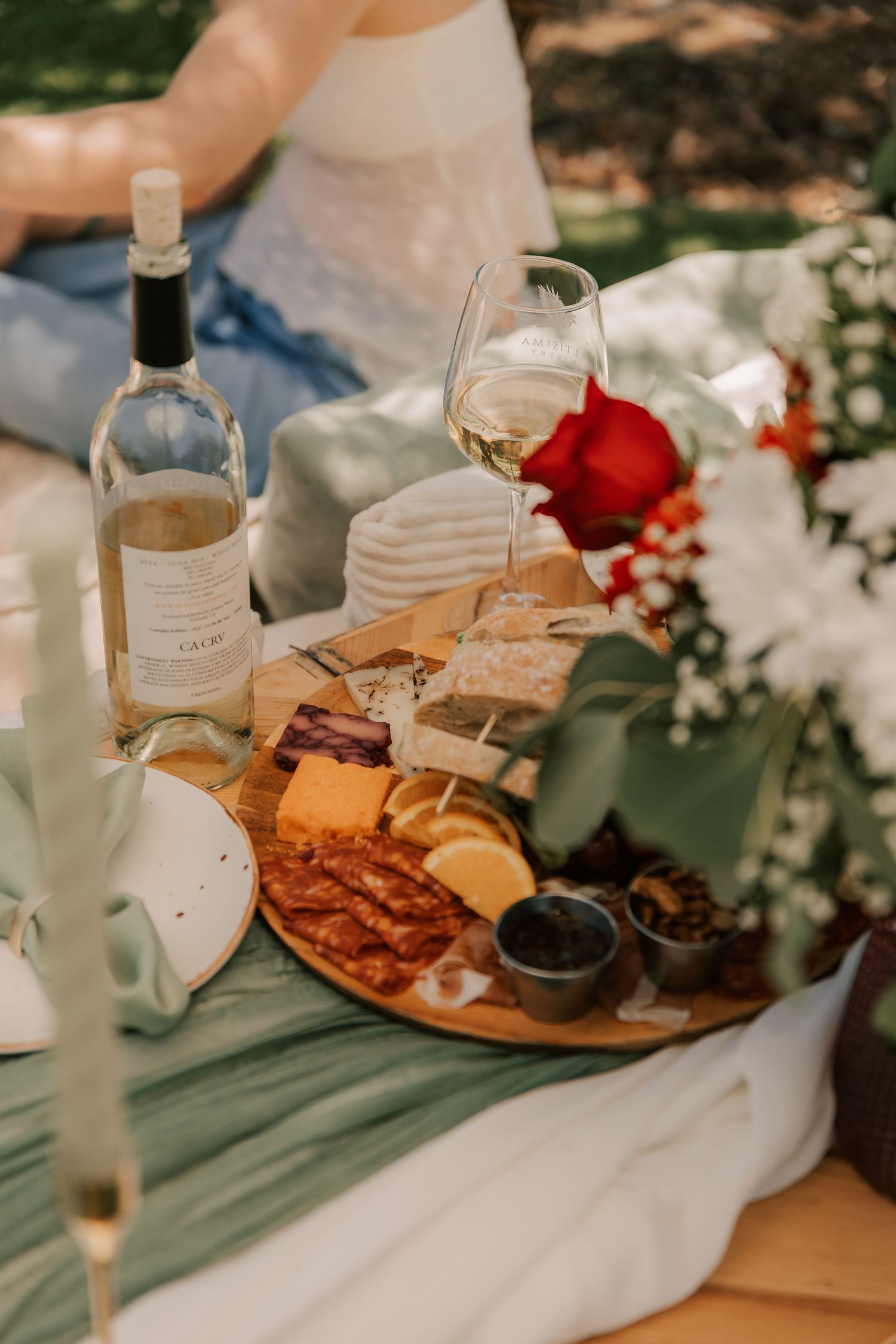 Elegant outdoor picnic with charcuterie board, white wine, cheese, and red rose on green blanket.
