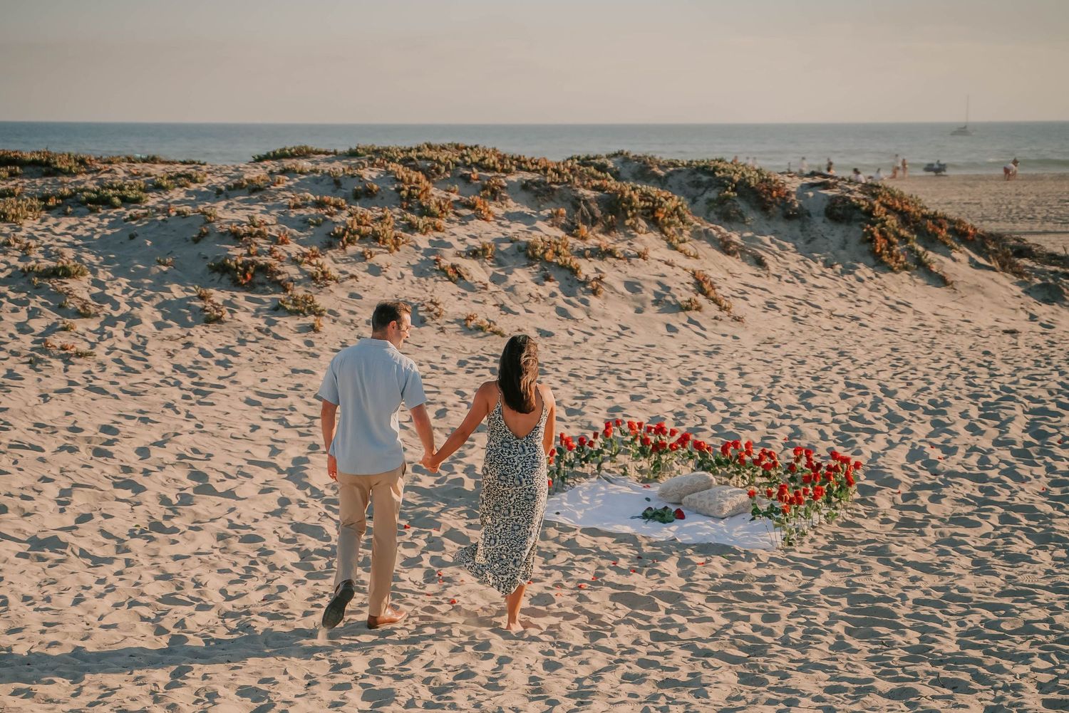 Couple holding hands walking toward a romantic floral arrangement on a sandy beach at golden hour sunset.
