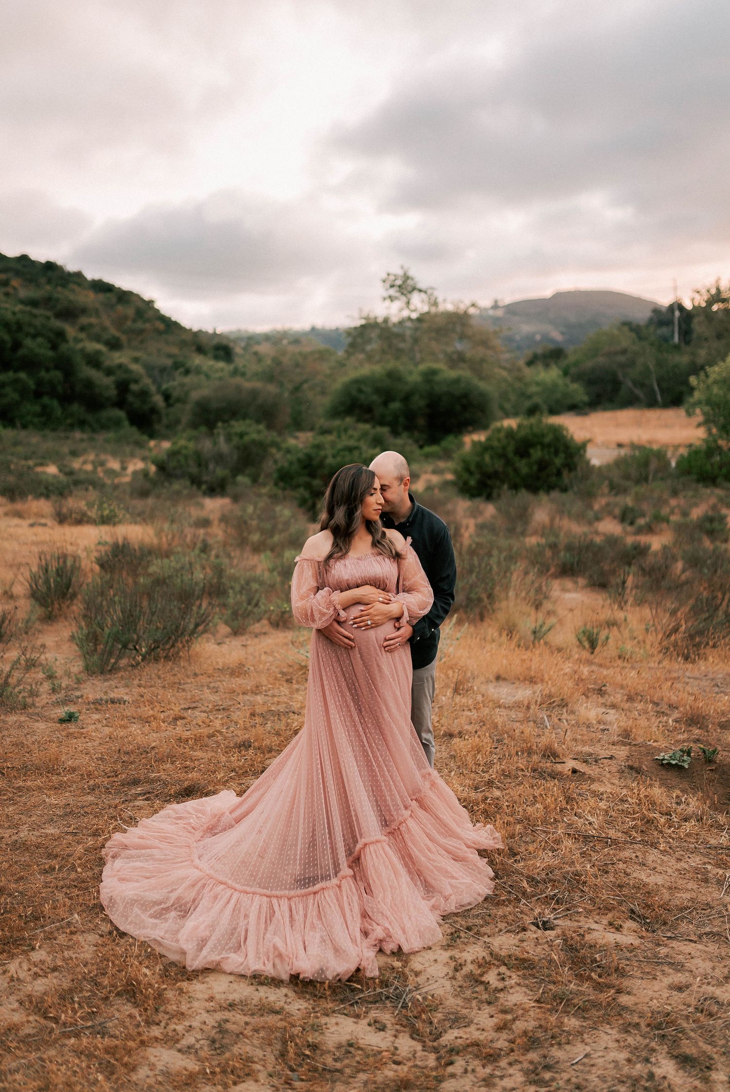 Pregnant woman in pink gown with partner in outdoor maternity photo shoot against scenic hills.