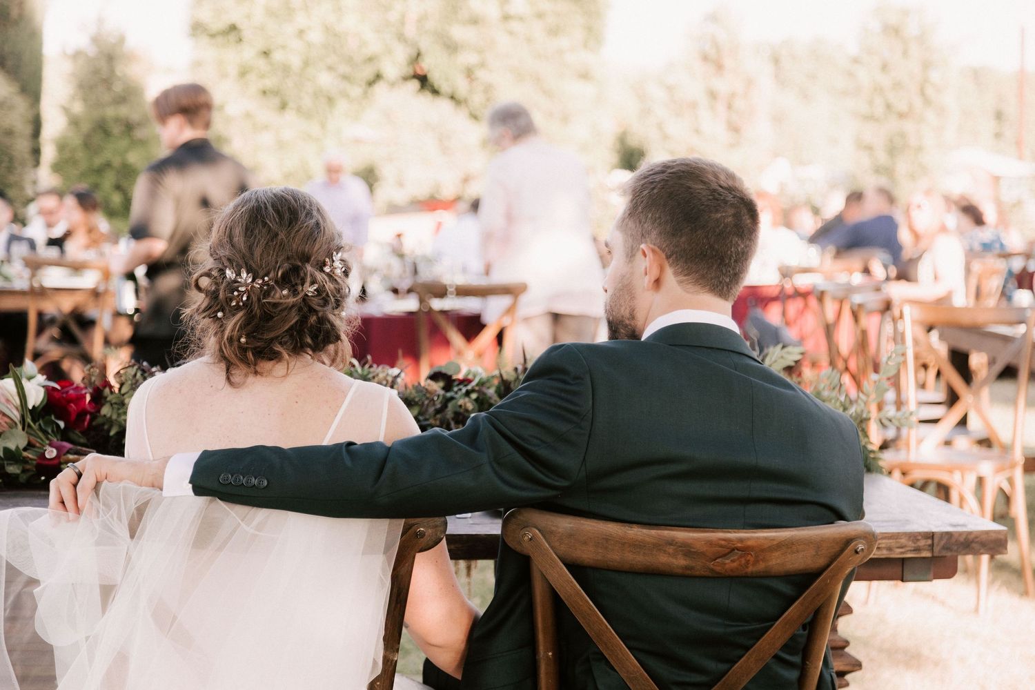 Bride and groom seated together at outdoor wedding reception, groom's arm around bride's chair.