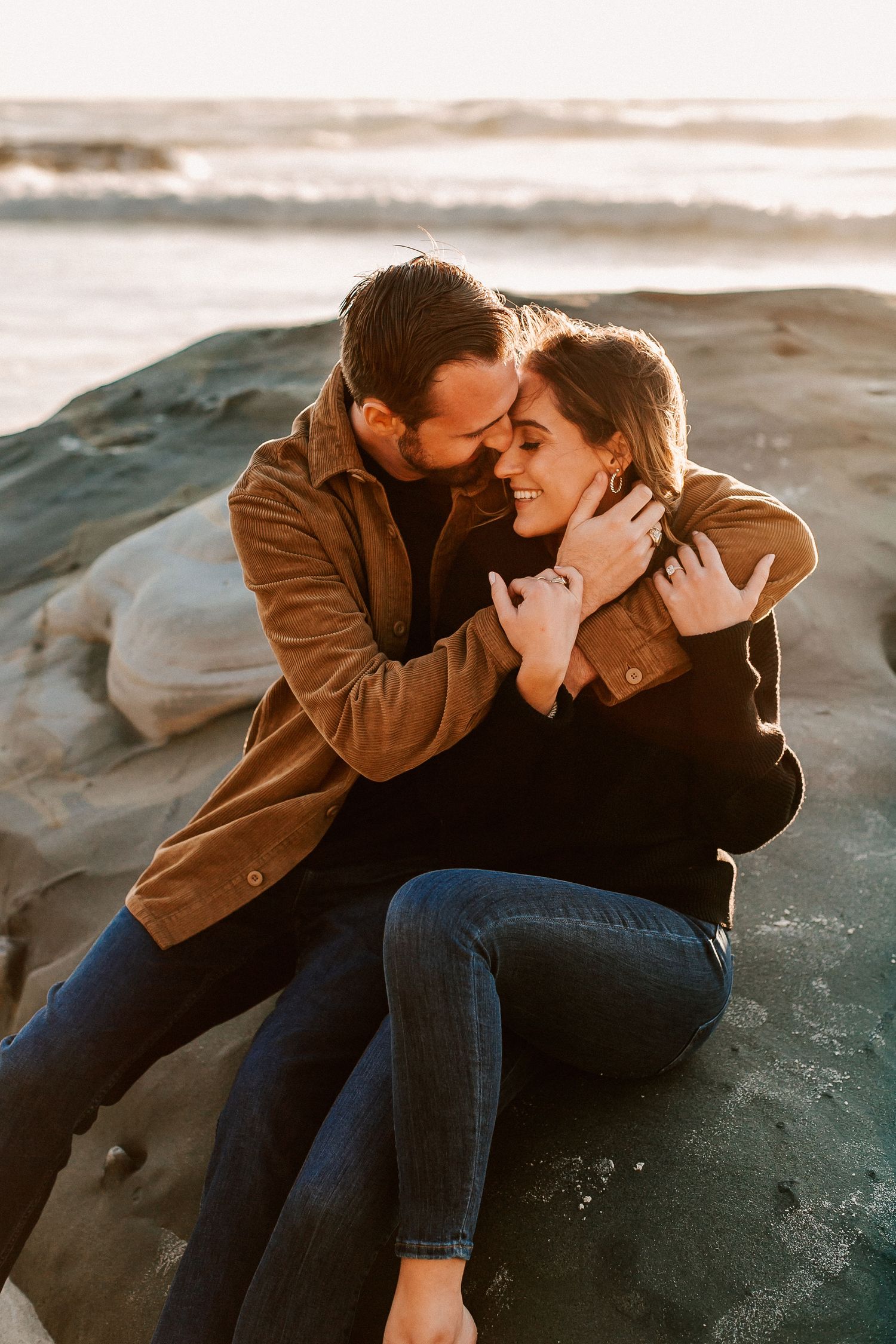 Couple sitting on beach rocks at sunset, smiling and embracing in warm golden light.