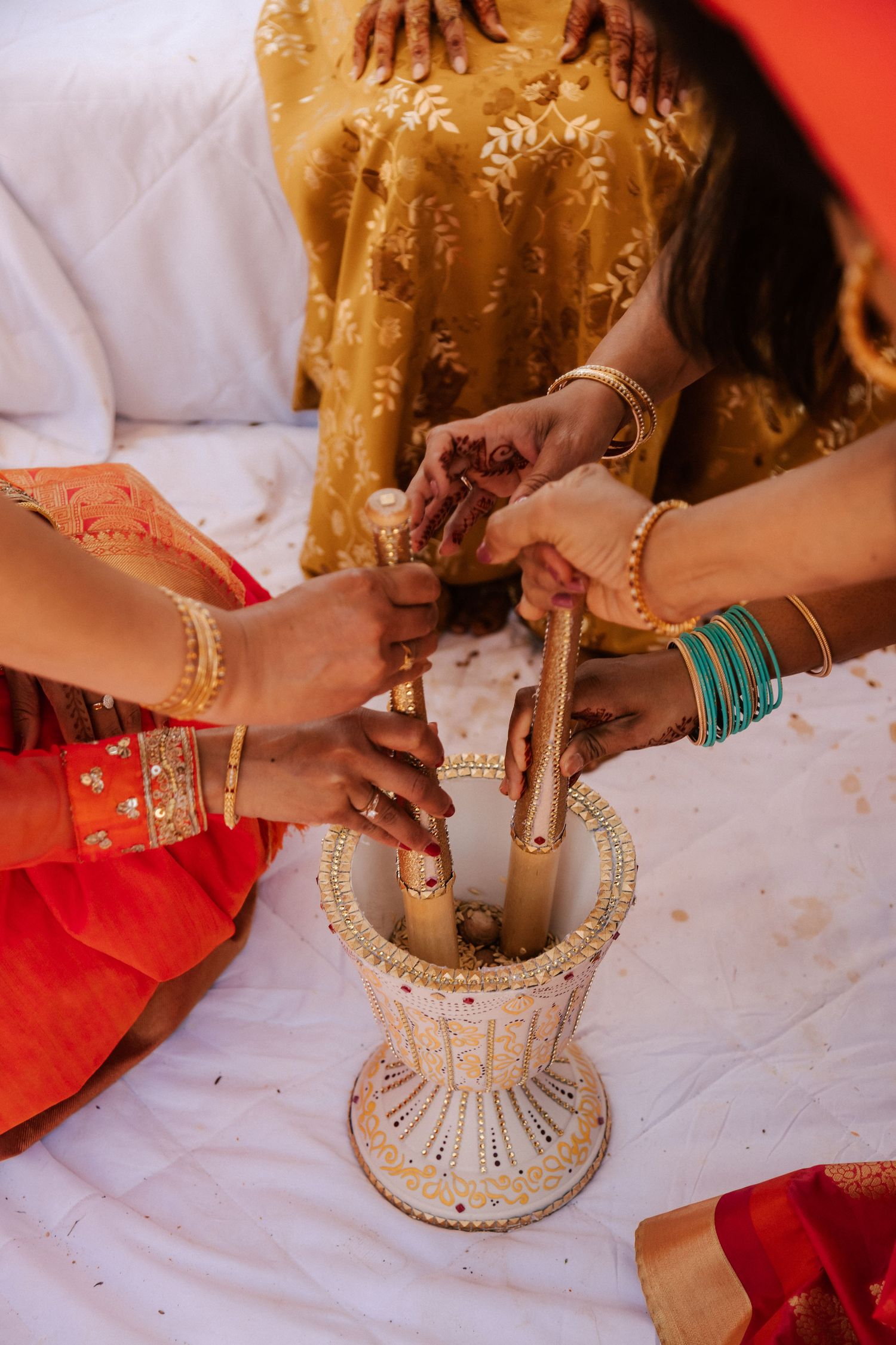 Multiple hands adorned with bangles grinding ingredients in decorated mortar during Indian wedding ceremony.