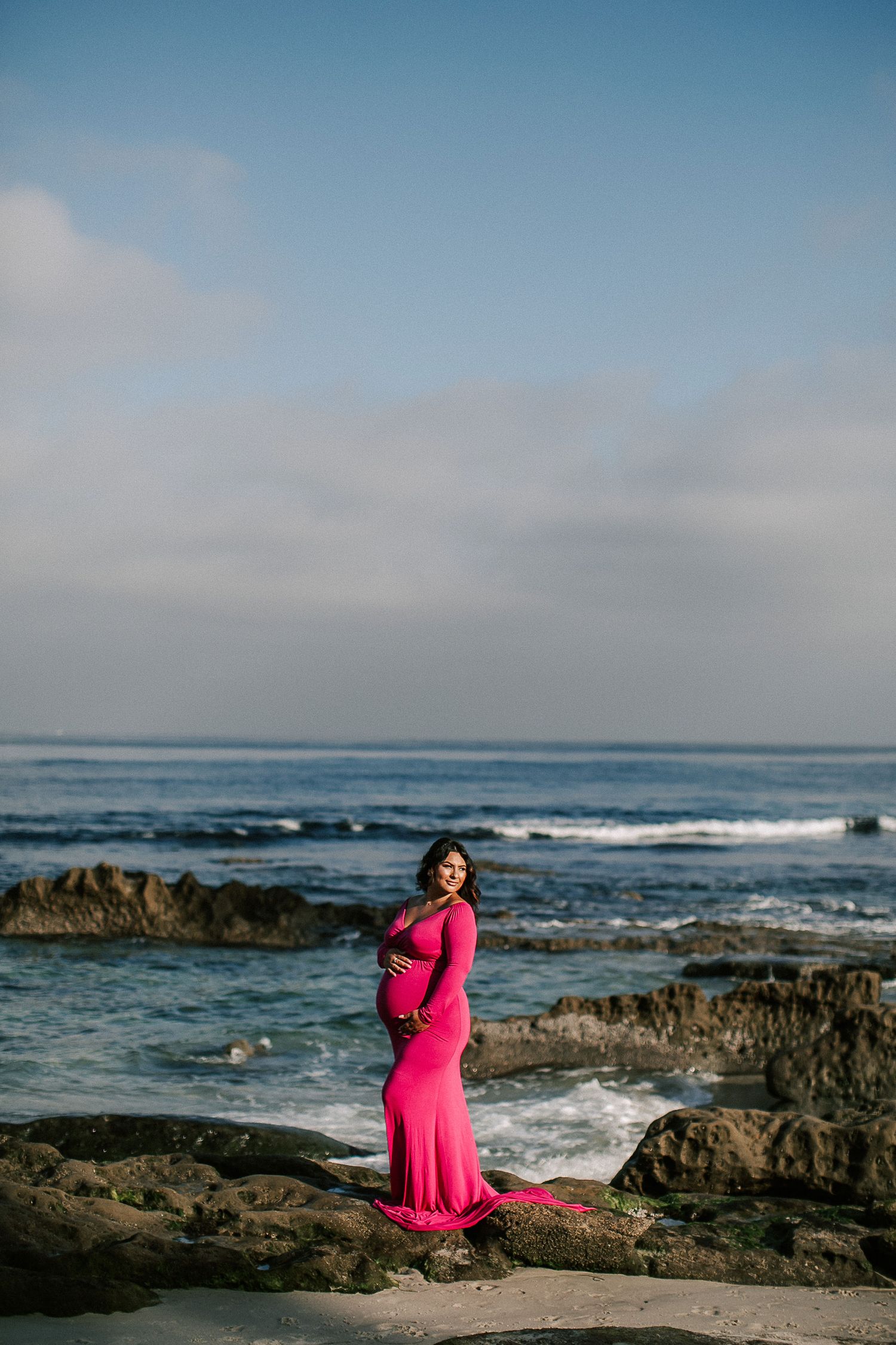 Pregnant woman in fitted hot pink maternity gown standing on rocky beach with ocean backdrop.
