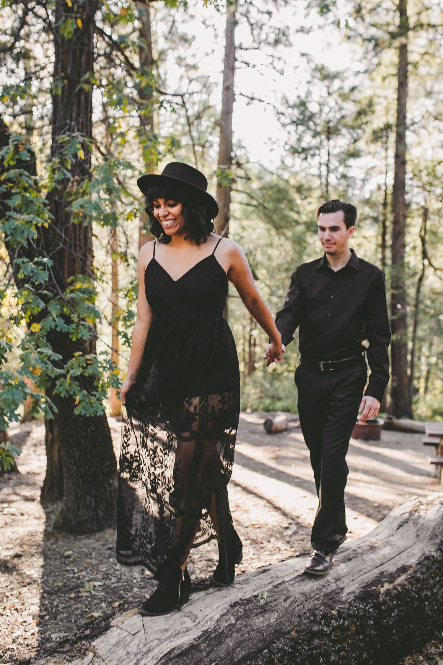 Couple in matching black outfits walking hand-in-hand on a log in a sunlit forest, woman wearing a hat and lace dress.