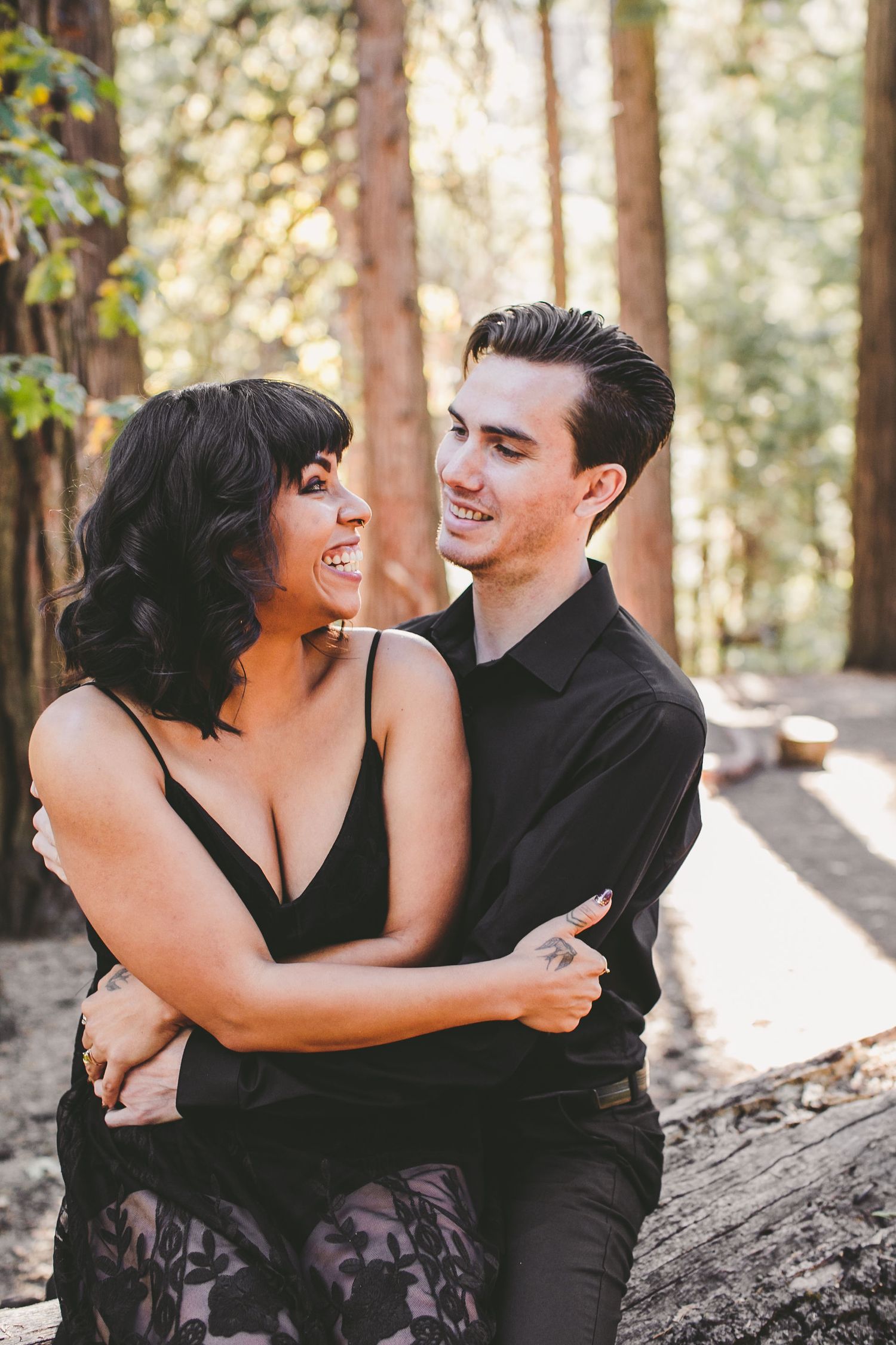 A smiling couple embraces romantically among tall trees in a sunlit forest, both dressed in black.