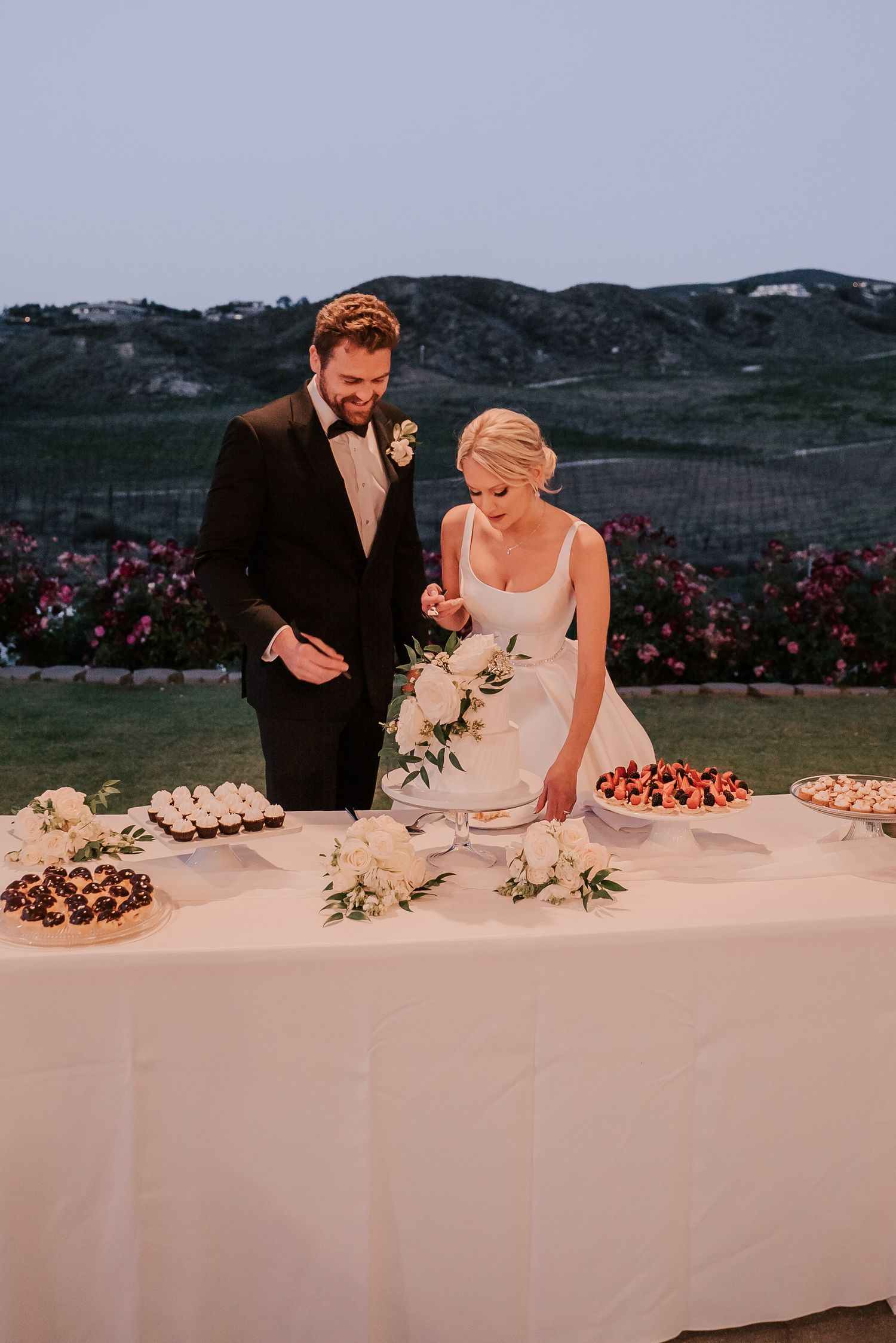 Bride and groom cutting wedding cake at outdoor reception table with desserts, flowers, and vineyard backdrop at dusk.