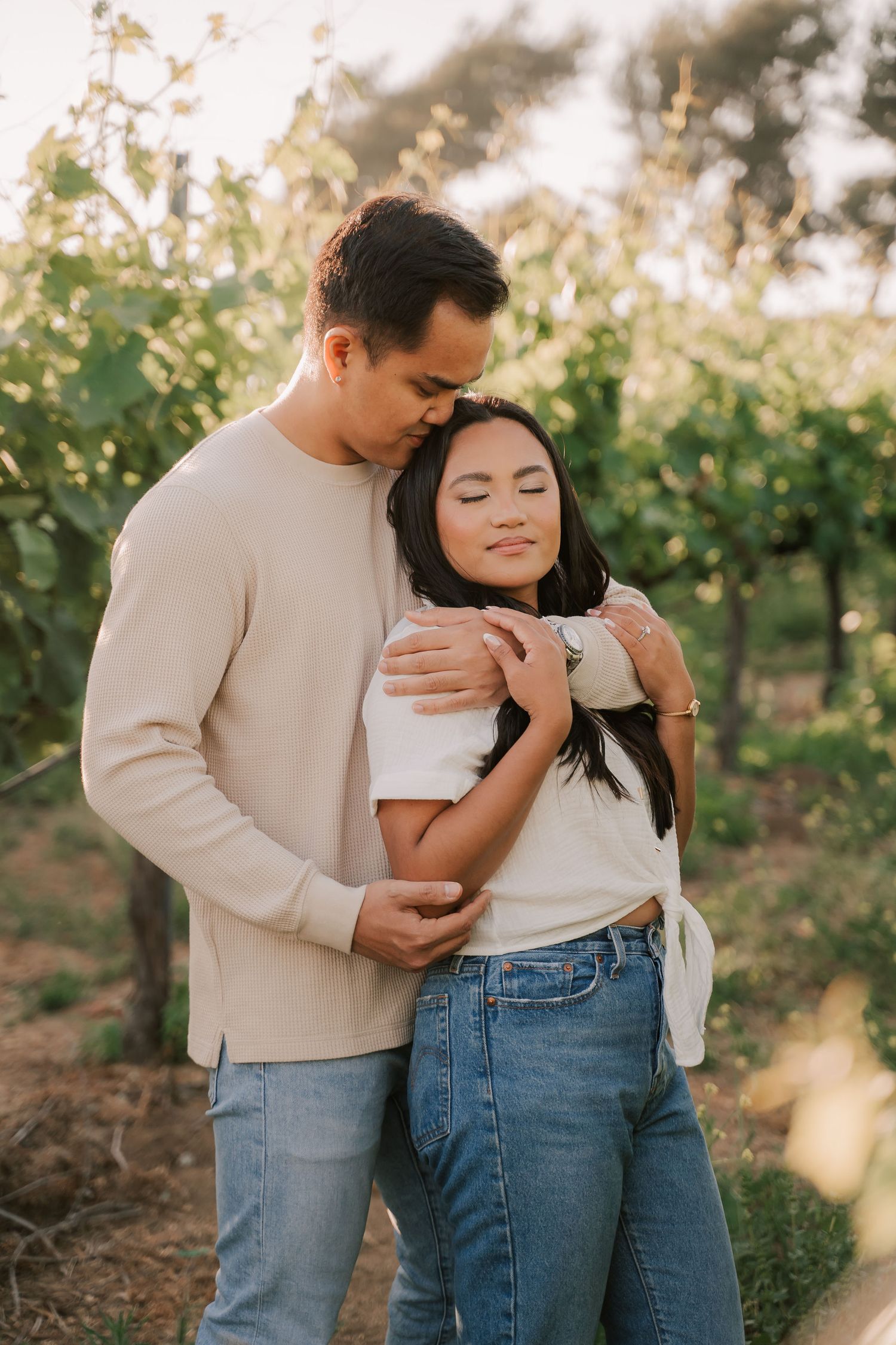 A couple embraces tenderly in a sunlit vineyard, the man kissing her head as she smiles peacefully in jeans and white top.
