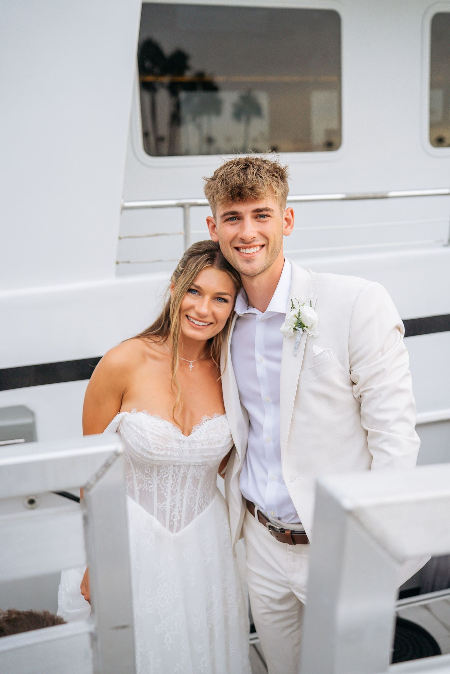 Smiling bride and groom pose together on a yacht, groom wearing a white suit with a floral boutonniere.