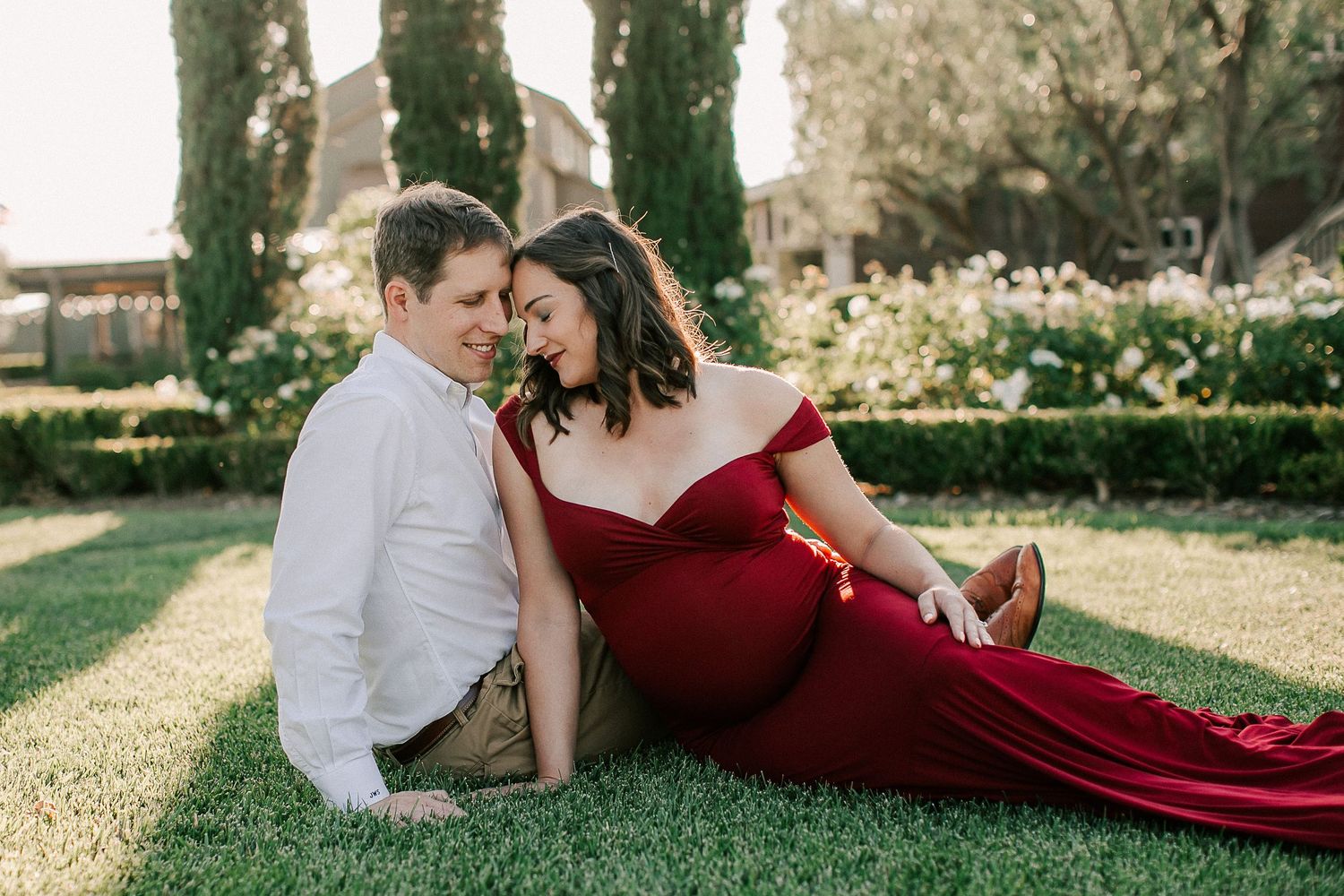 Couple in maternity photoshoot sitting on grass, woman in red off-shoulder gown, man in white shirt, garden setting.