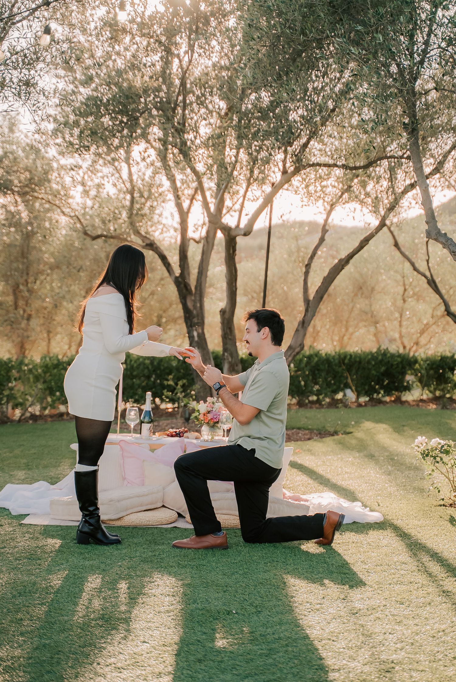 Man kneeling on one knee proposing to woman at romantic outdoor picnic setup surrounded by trees at golden hour.