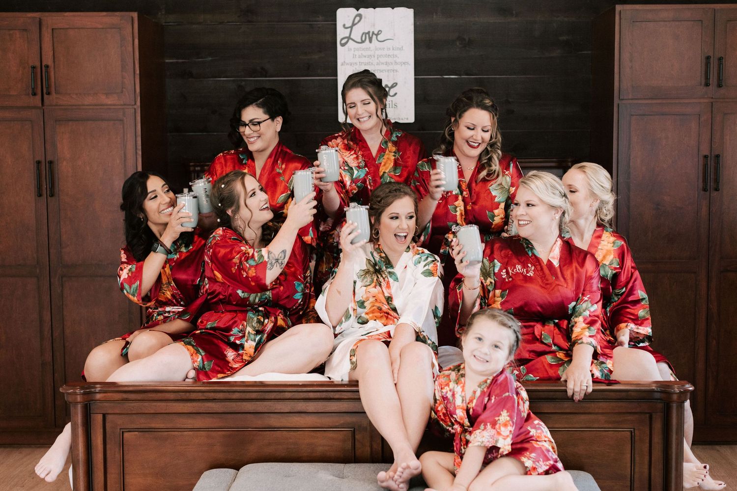 Bridesmaids in red floral robes laughing together on a bed, holding champagne glasses, with a flower girl seated in front.