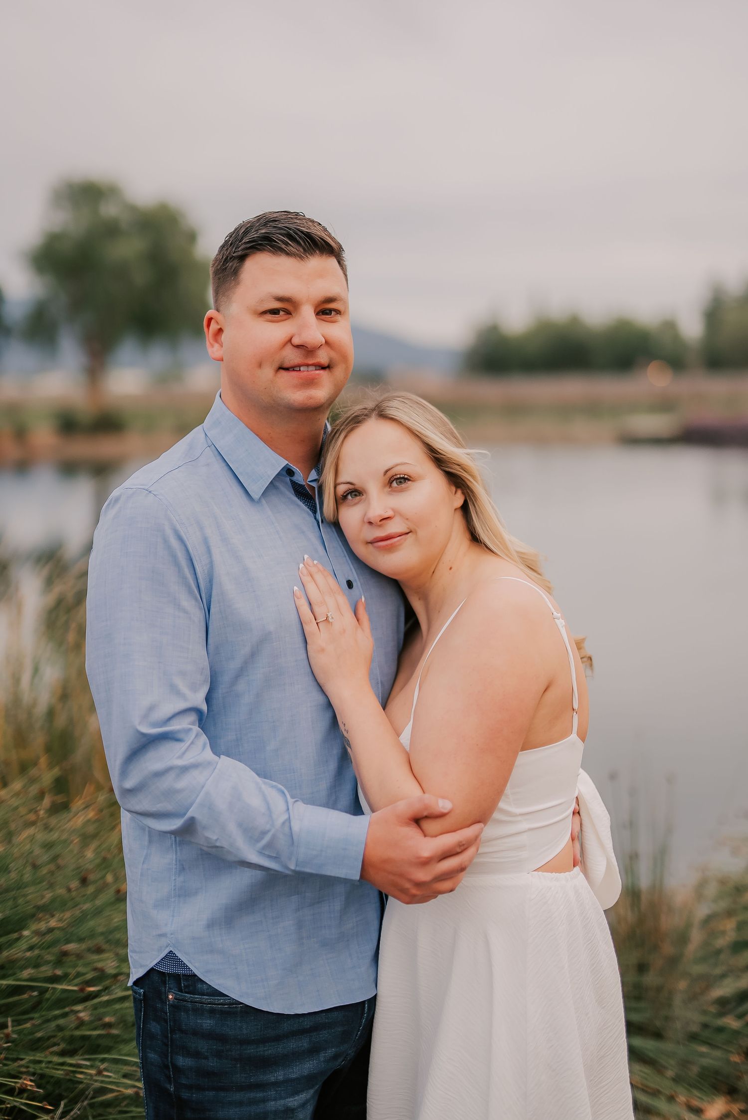 Couple embracing outdoors near a lake at golden hour, woman in white dress and man in blue shirt smiling at camera.