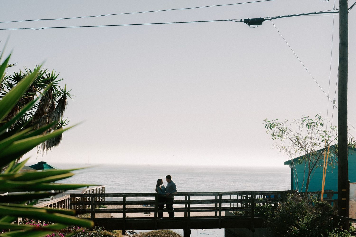 Couple silhouetted on a wooden bridge overlooking the ocean at dusk, surrounded by tropical plants.