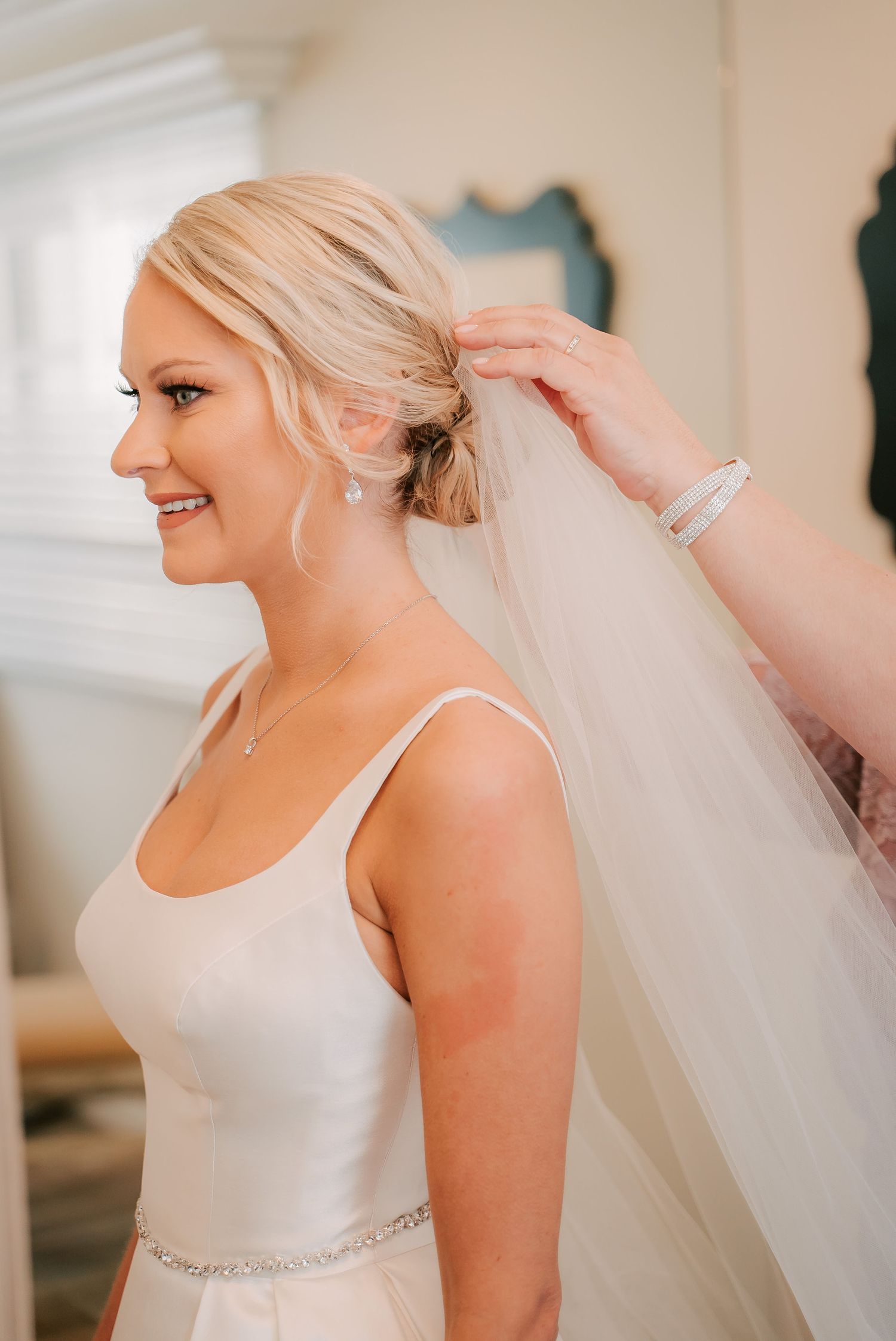 Bride smiling in white gown with veil being pinned into updo hairstyle, wearing pearl earrings and necklace.