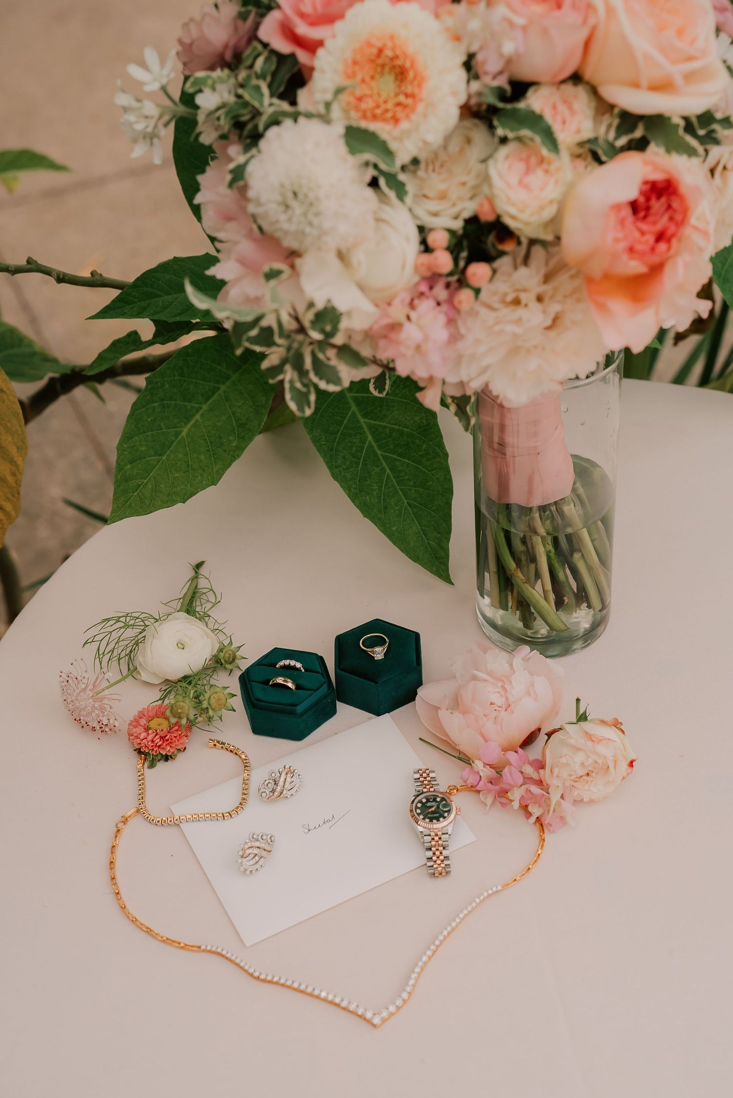 Elegant wedding flat lay with green velvet ring boxes, pearl necklace, Rolex watch, earrings, and pink floral bouquet on white table.