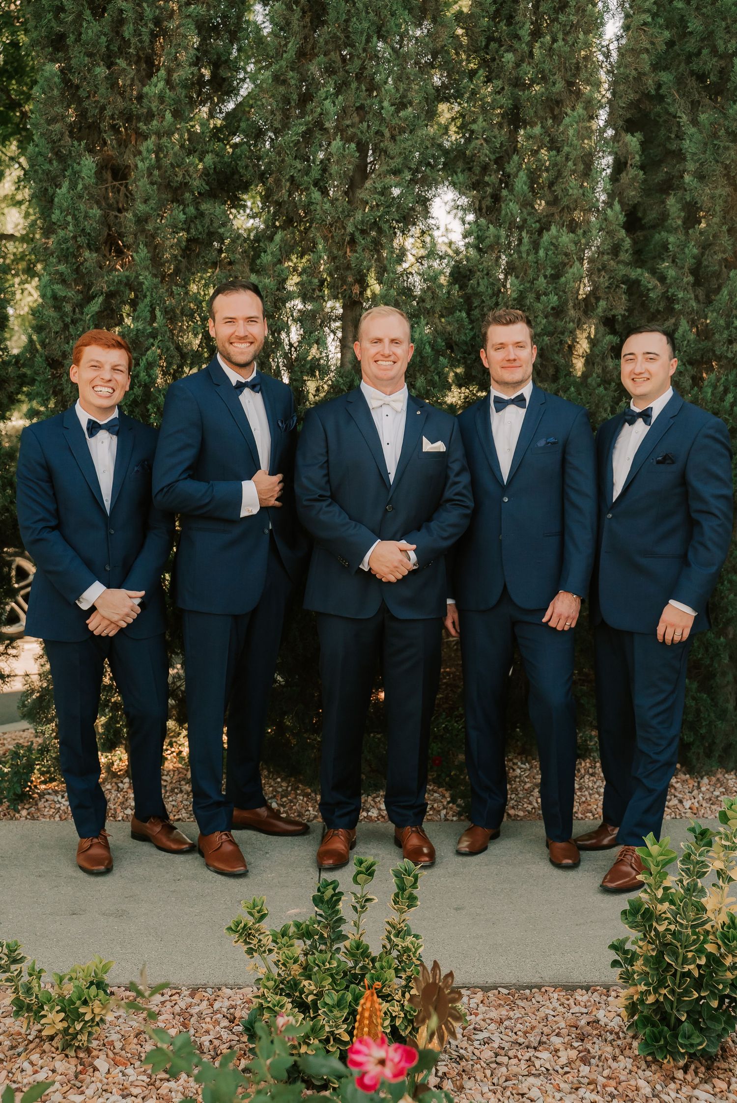 Five groomsmen in navy blue suits and bow ties pose together outdoors near lush green trees at a wedding.