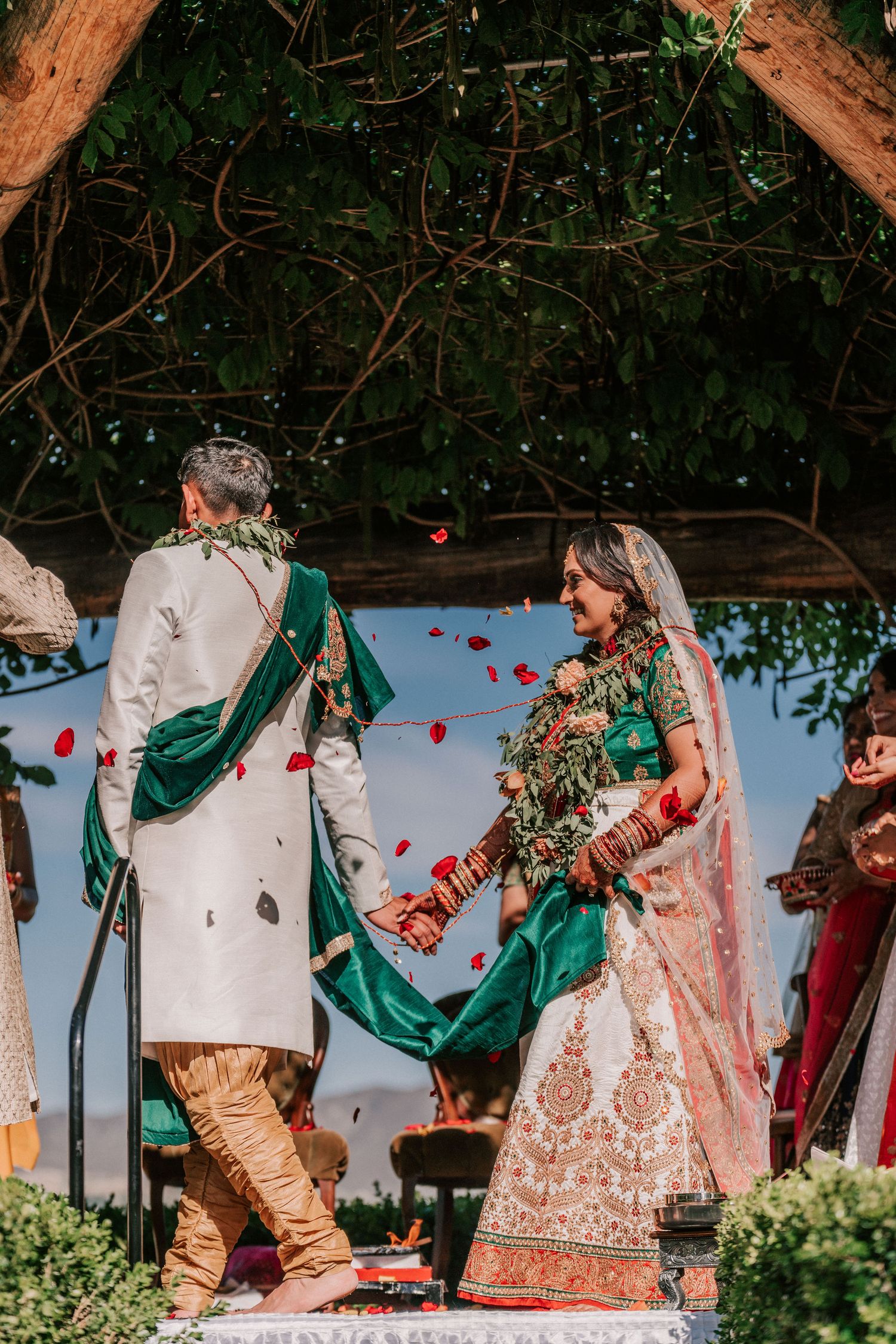 Indian bride and groom holding hands as rose petals fall during outdoor ceremony.