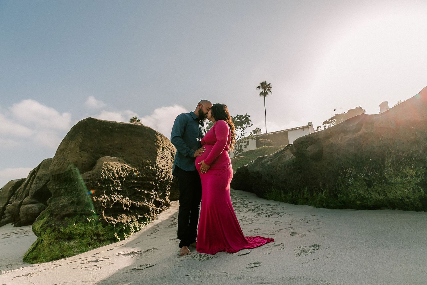 Pregnant couple sharing a tender moment on sandy beach with rocks and palm tree in golden sunlight.