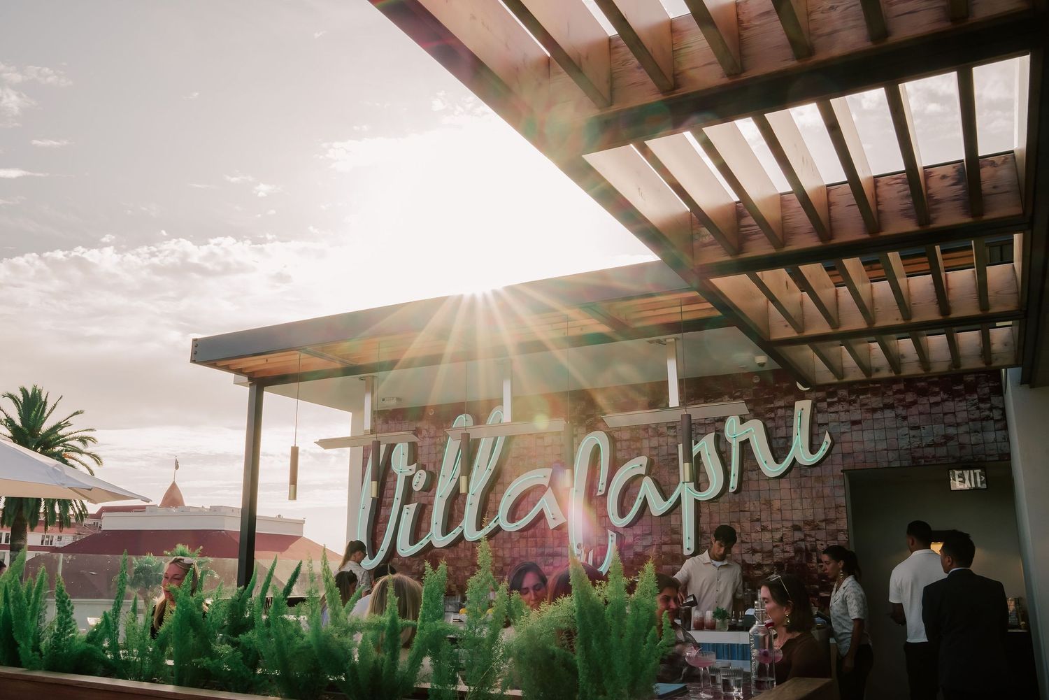 Sunlit Villa Capri sign with lush greenery, brick wall, and guests gathered on a rooftop terrace at golden hour.