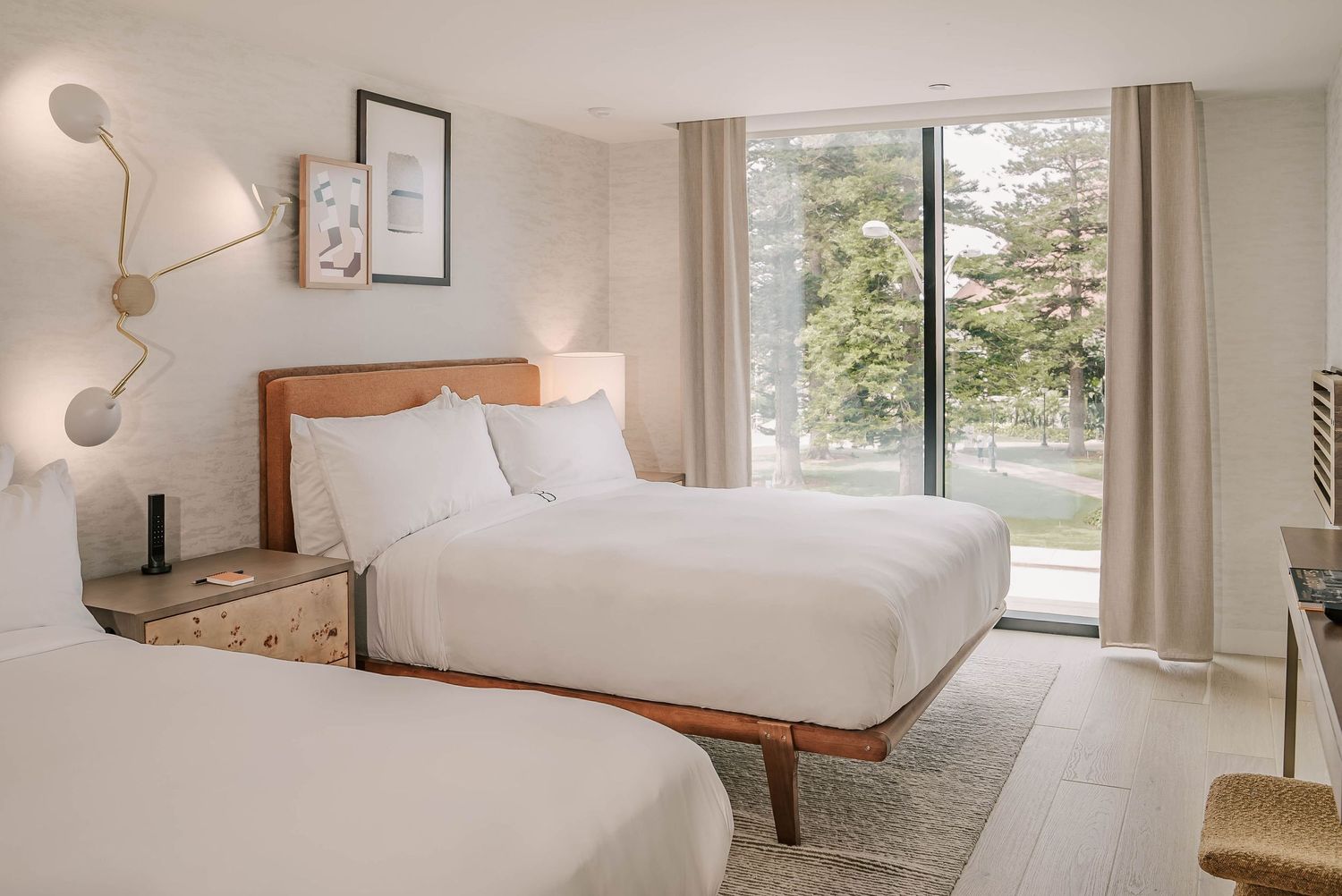Bright hotel room with two queen beds, white bedding, wood frames, neutral decor, and large window with garden view.