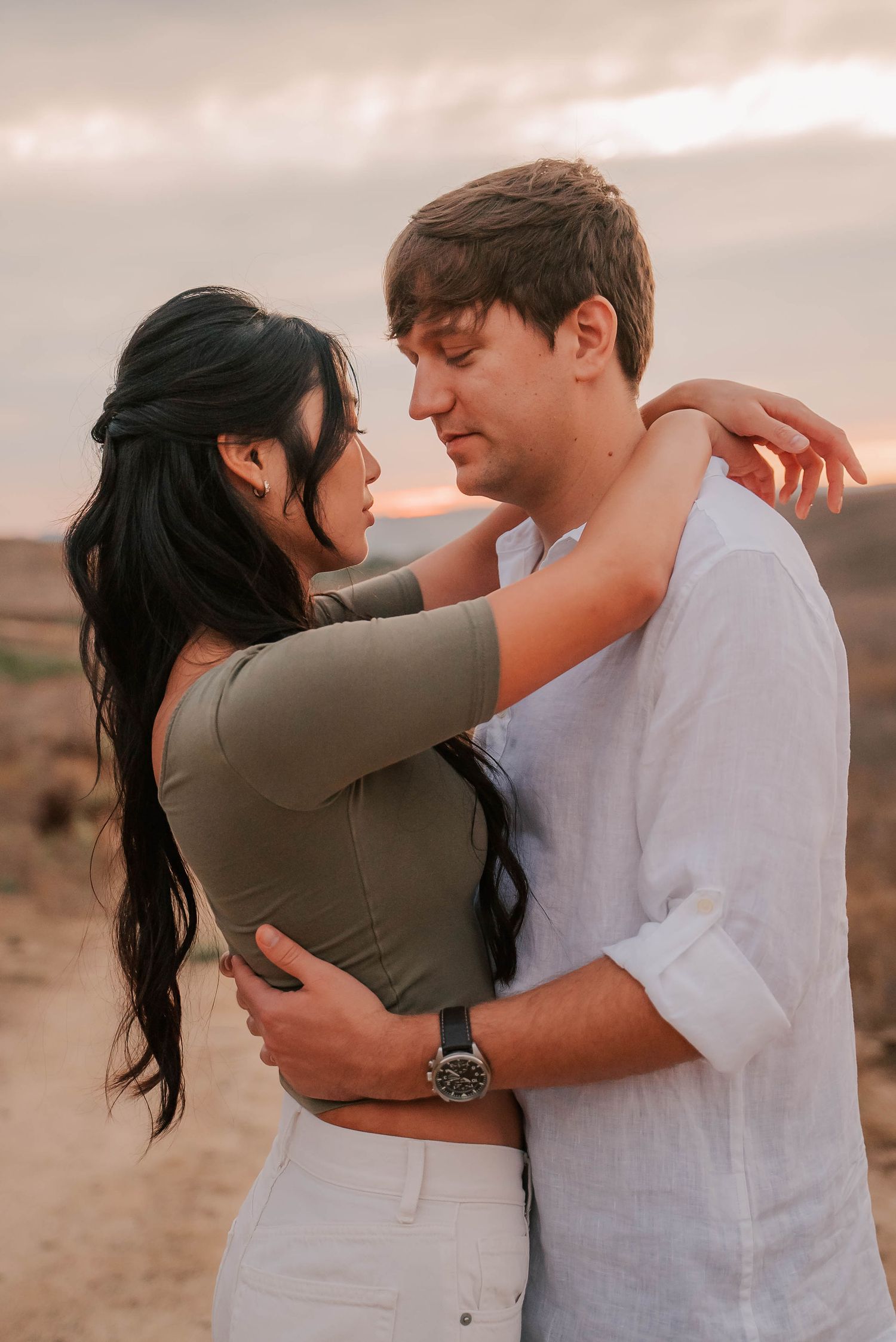 Couple embracing at sunset in desert, woman in olive top with long dark hair, man in white linen shirt wearing a watch.