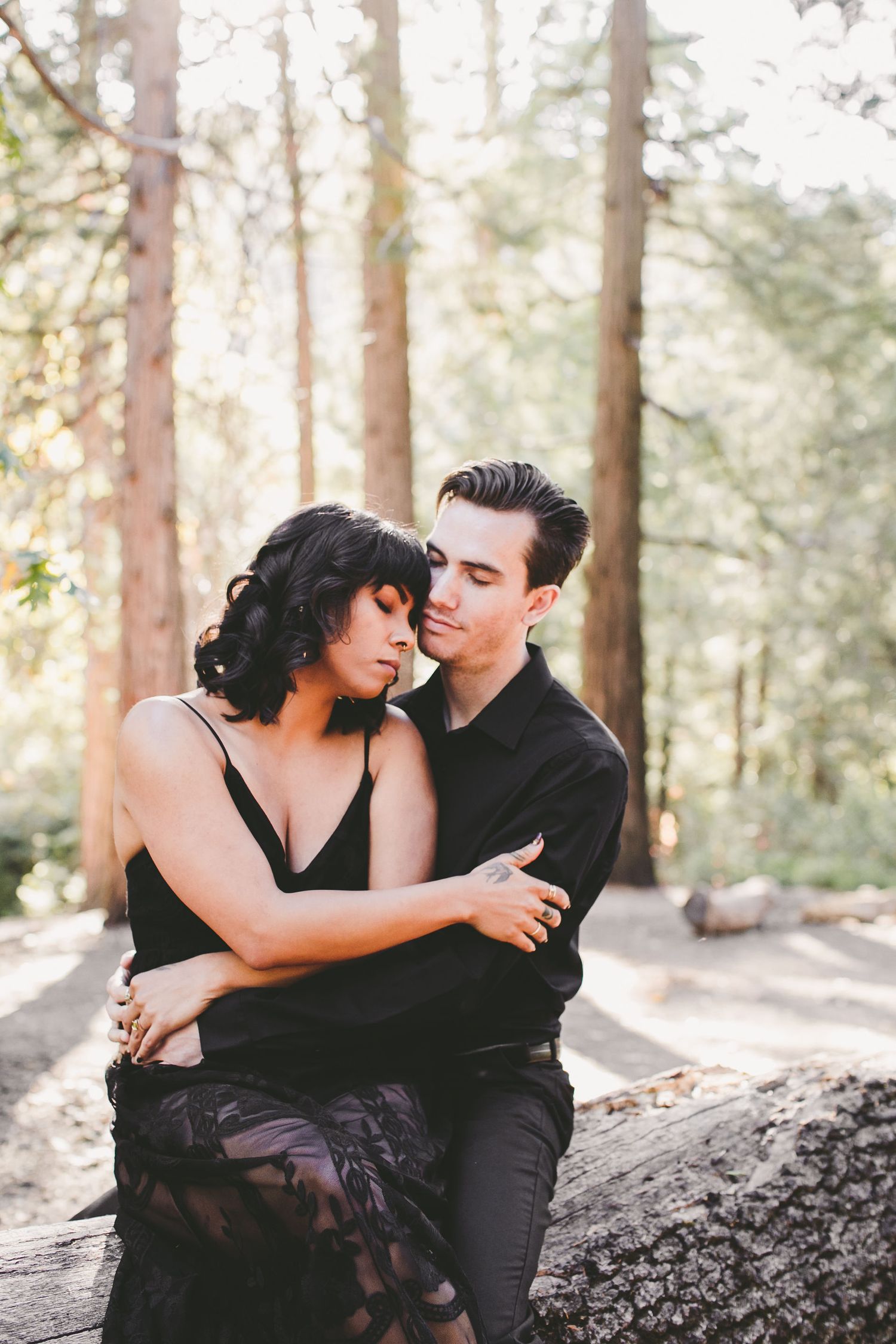 Couple in black outfits embracing tenderly on a log in a sunlit forest setting.