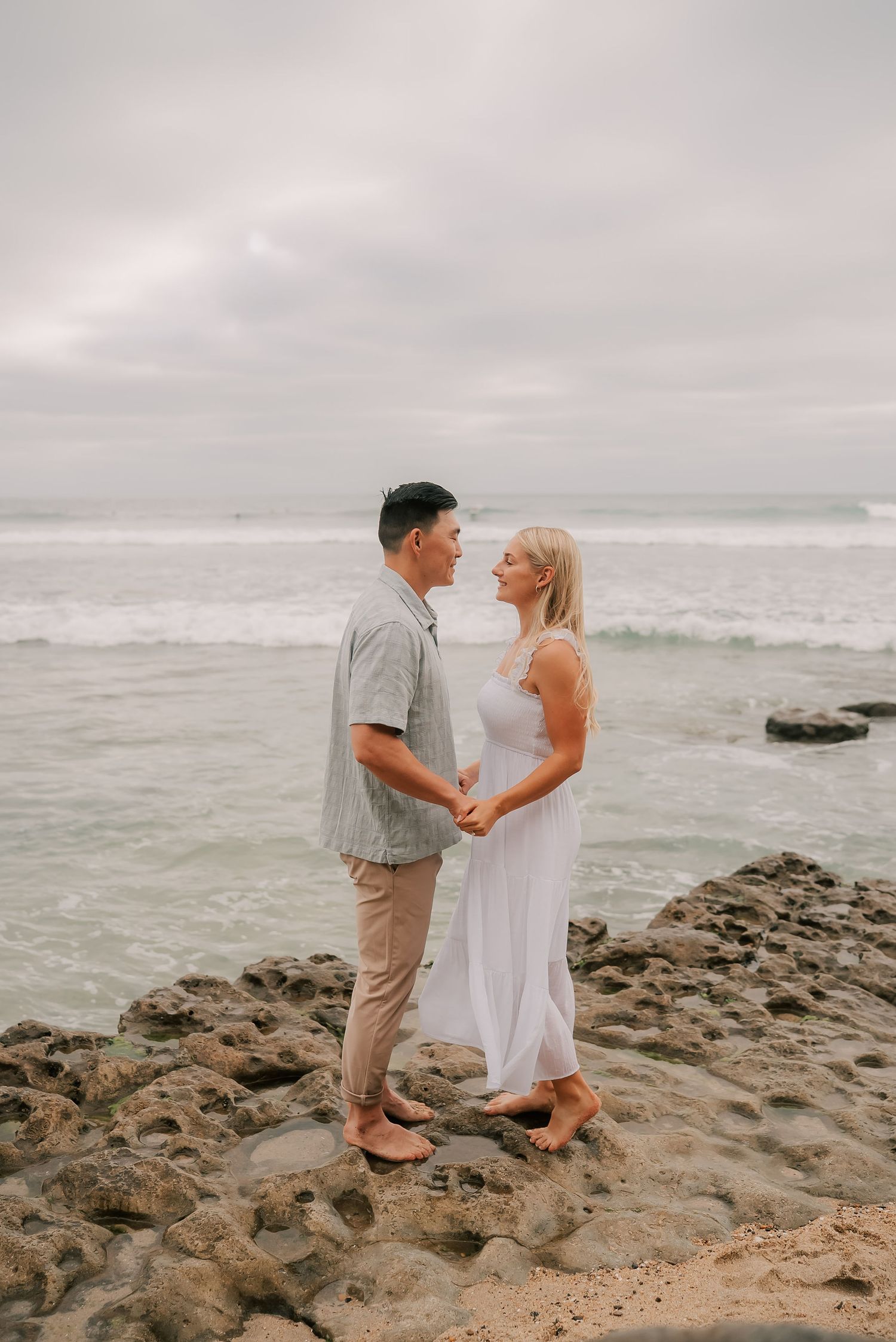 Couple holding hands on rocky beach shore, wearing white dress and gray shirt, with ocean waves in background.