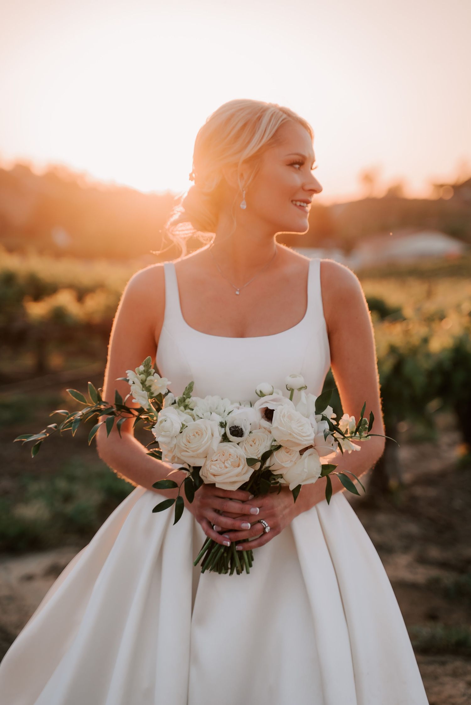 Smiling bride in white gown holding white rose bouquet at golden hour in vineyard.