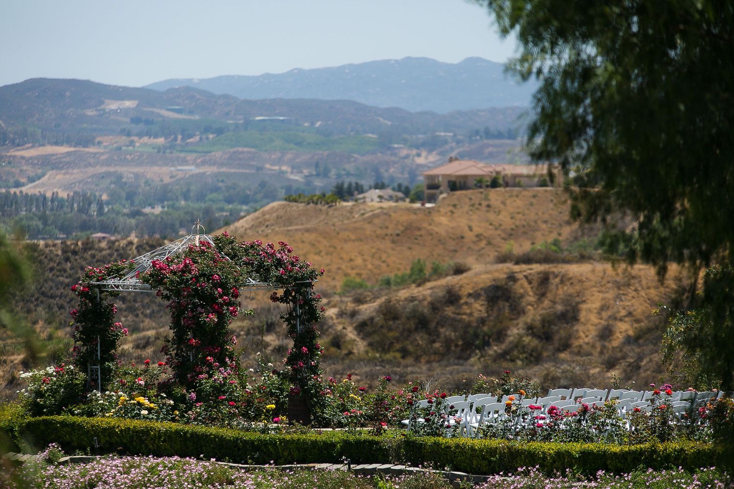 Rose-covered garden arches overlook a scenic valley with rolling hills and colorful flower beds under a clear blue sky.
