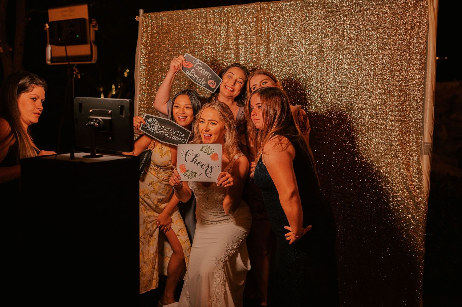 Bride and bridesmaids posing with fun photo booth props in front of a gold sequin backdrop at a wedding reception.