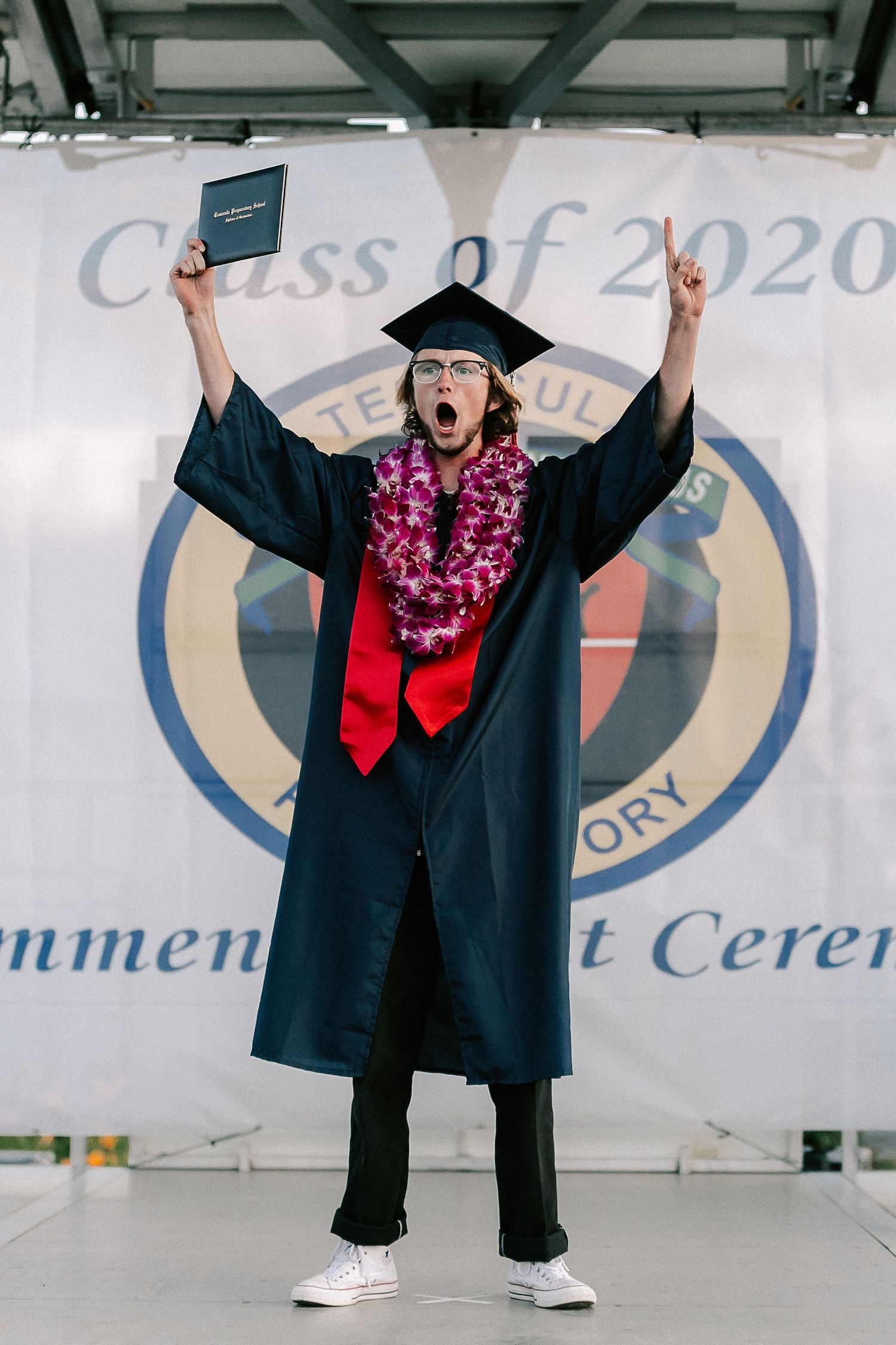 Excited Class of 2020 graduate in cap and gown raises diploma and finger at commencement ceremony.
