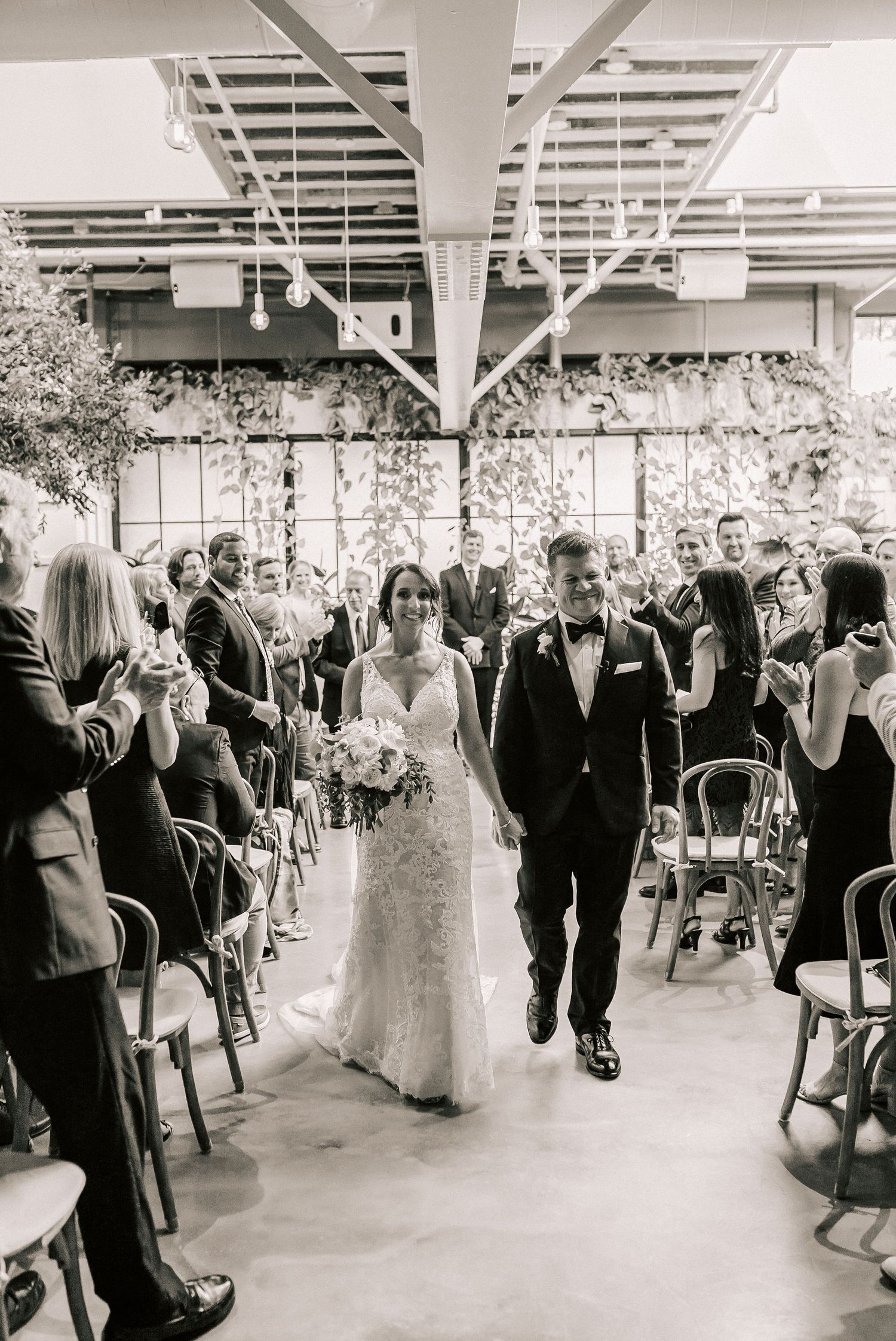 Bride and groom walk down the aisle together at an elegant indoor wedding ceremony with lush greenery decor.