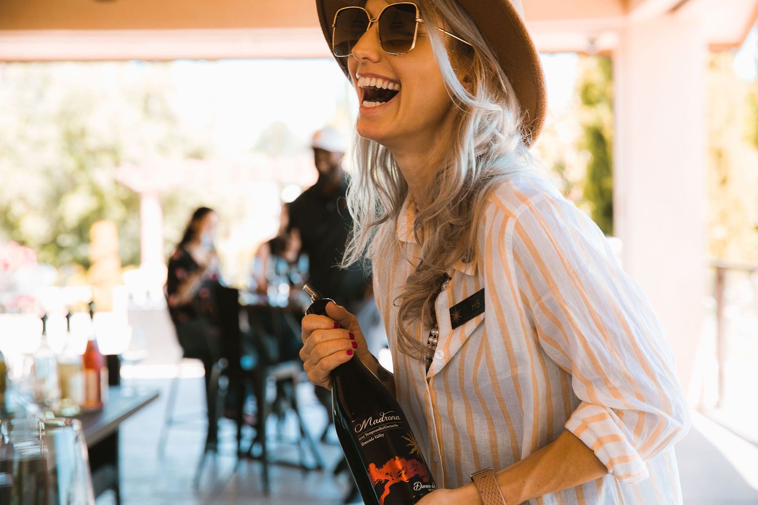 Smiling woman in sunglasses and hat opening a champagne bottle at an outdoor venue.