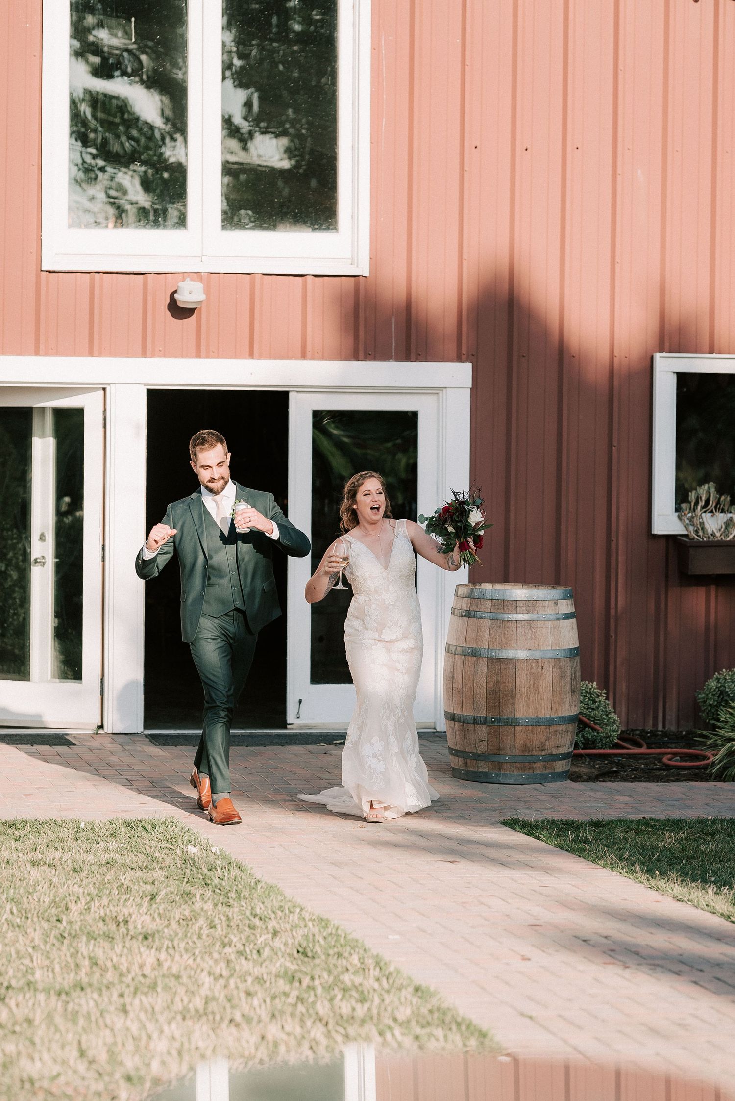 Joyful bride and groom exit a rustic red barn venue, celebrating their wedding day with bouquet in hand.