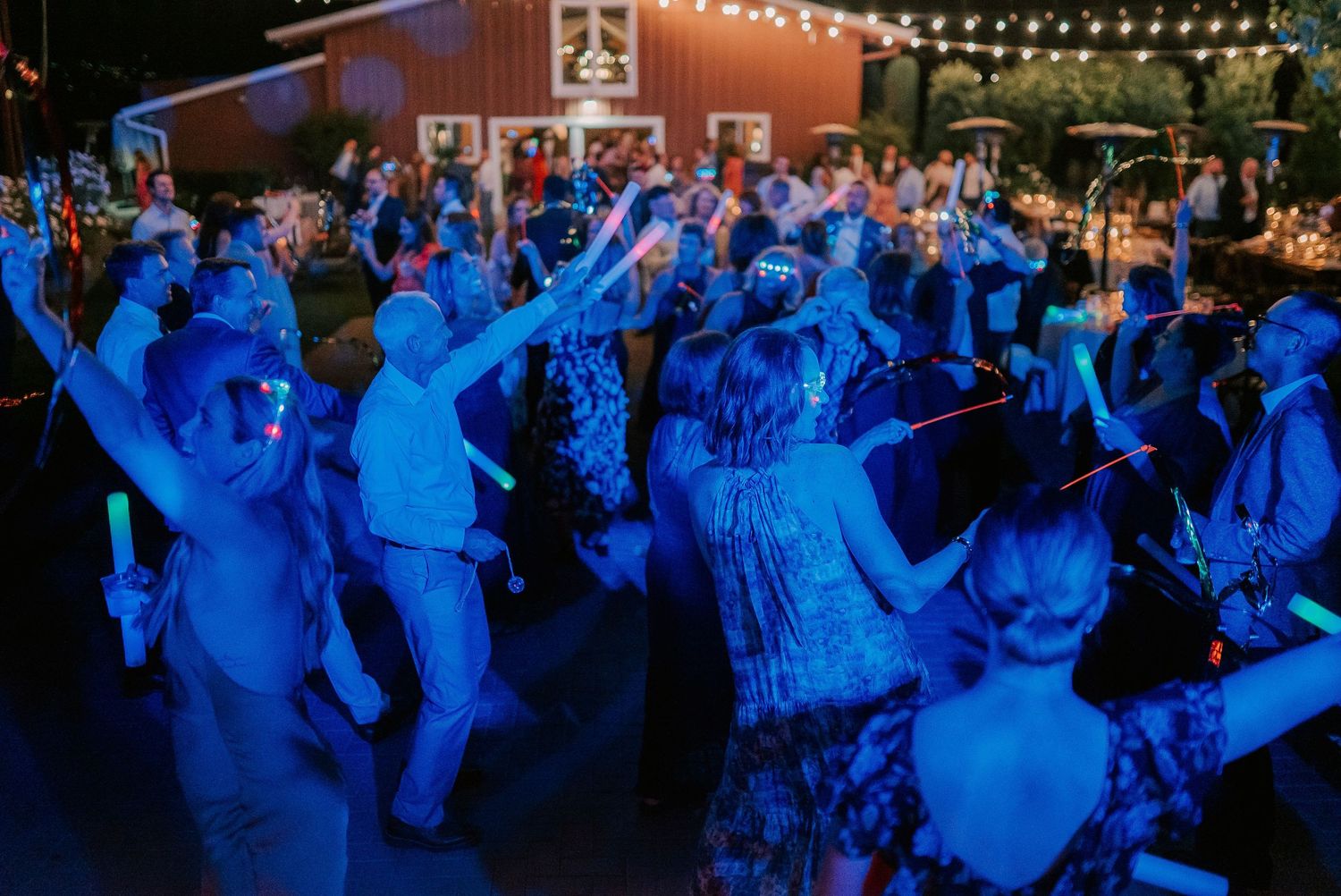 Guests dancing under blue lights at an outdoor wedding reception near a red barn at night.