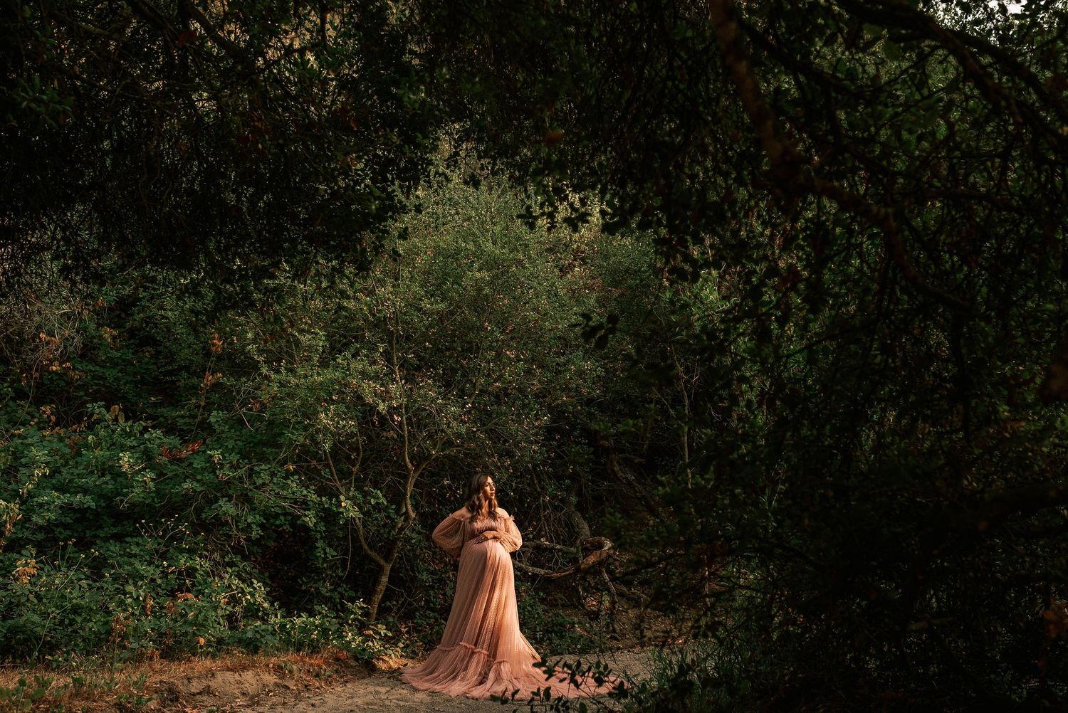Woman in flowing peach gown standing in lush green forest, surrounded by trees in a dreamy outdoor portrait.
